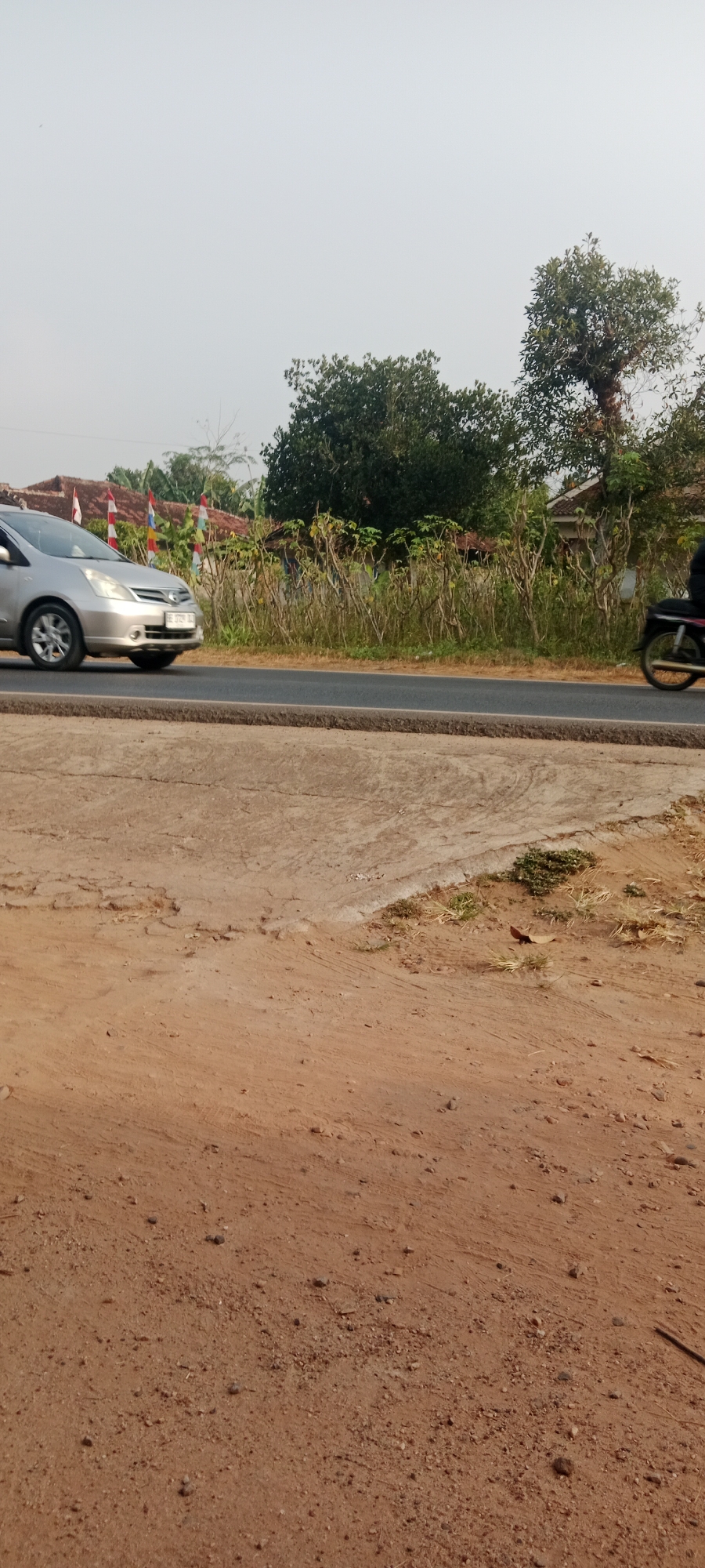 The image captures a roadside scene in Gondang Rejo, Indonesia. A silver car is moving along a paved road, which is bordered by a dirt and gravel shoulder in the foreground. Behind the road, a line of dry vegetation and small trees separates the road from a row of houses with tiled roofs. Several Indonesian flags are visible in front of the houses, indicating a festive or commemorative occasion. In the background, lush green trees and a larger, mature tree with a thick trunk stand against a hazy sky. On the right side of the road, a person on a motorcycle is partially visible, also in motion. The overall impression is a typical village or suburban street in Indonesia.