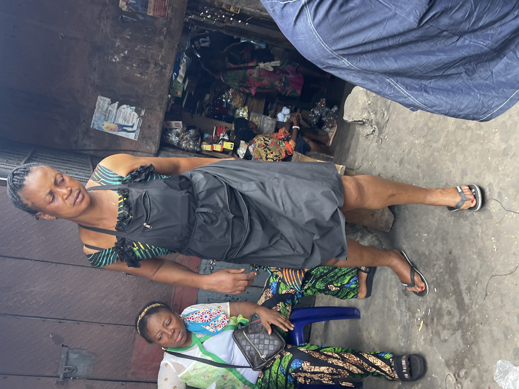 The image depicts a street scene in Nkpor, Nigeria. A woman in a black dress stands with another woman seated on a blue plastic chair. The seated woman is wearing a patterned dress with white top. A handbag rests on her lap. Both are wearing sandals. The background reveals a cluttered market stall with various goods on display. A metal corrugated gate is visible to the side. Overhead, a blue tarp is partially visible, suggesting a covered market area. The ground is grey concrete.