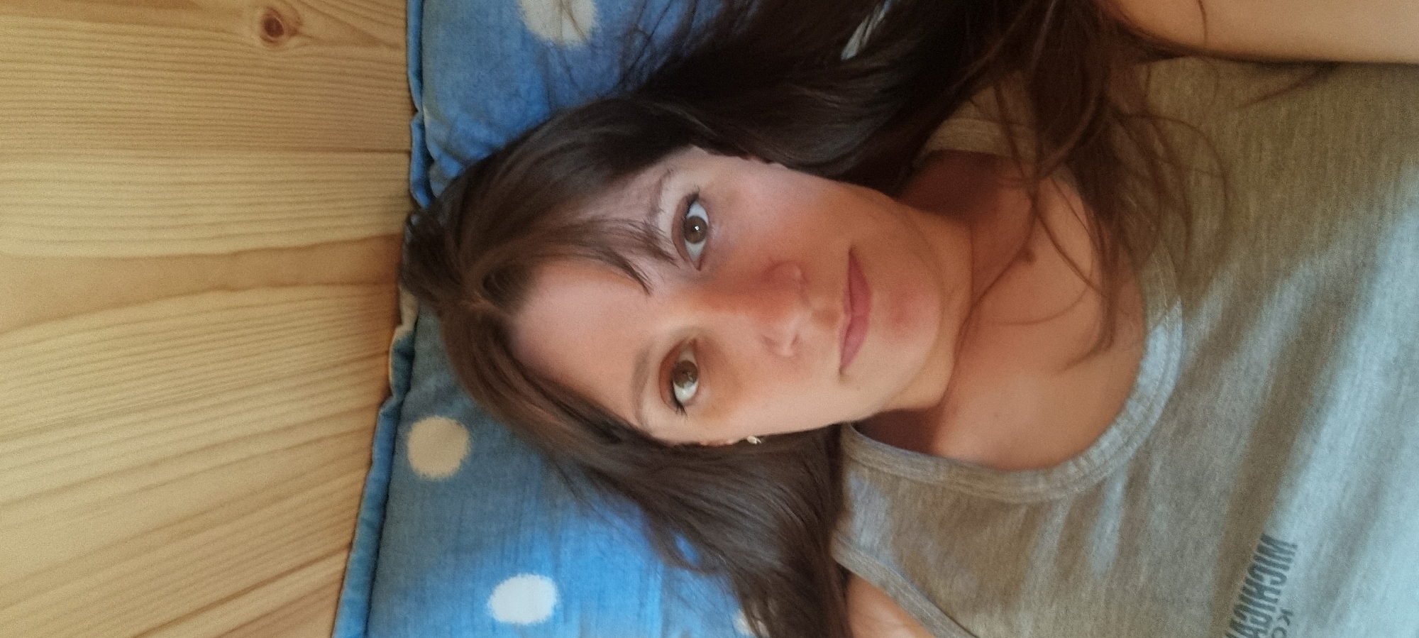 The image, taken in Dunnae-myeon, South Korea, captures a young woman lying down and looking directly at the camera. The shot is oriented vertically, with her head towards the top of the frame. She has long, dark brown hair with bangs across her forehead, and warm brown eyes. Her expression is calm and neutral.
She is wearing a plain grey t-shirt. On the front of the shirt, reversed and partially visible, there is white text that appears to spell out elements like "WICHICK" or "MICHIGA" and "CO" below, possibly indicating a brand or place name like "MICHIGAN CO" or "CHICAGO CO" when un-mirrored.
Her head rests on a blue pillow featuring white polka dots. To the left side of the frame, a background of light-colored wooden planks is visible, likely a floor or wall paneling. The lighting is soft and natural, suggesting an indoor setting during the daytime. The overall scene conveys a sense of quiet relaxation.