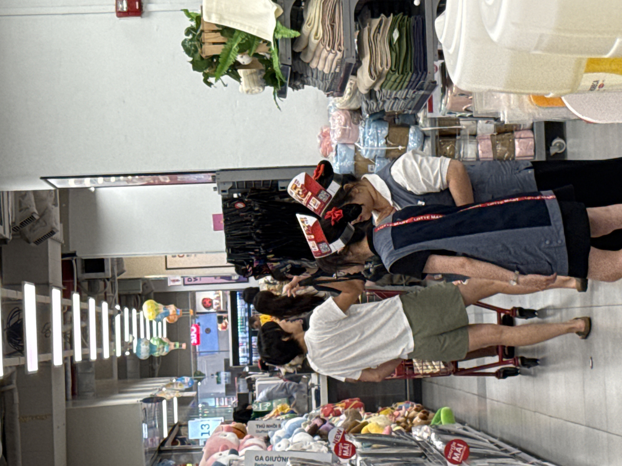 The image shows an interior view of a supermarket aisle in Nha Trang, Vietnam. In the foreground, a person wearing a white t-shirt and olive green shorts stands facing left, with a red shopping cart partially visible in front of them. To the right, two individuals are engaged in a conversation. One is wearing a blue vest over a white shirt and has decorative red and white headwear. The other, partially obscured, is wearing a dark vest. The aisle is lined with shelves displaying various merchandise, including plush toys on the left and stacks of folded textiles, possibly towels or blankets, on the right. Overhead, fluorescent lighting illuminates the scene. Colorful decorative elements, resembling hot air balloons, hang from the ceiling. A red fire alarm is visible on the wall to the far right. The overall environment suggests a retail setting with customers and staff.