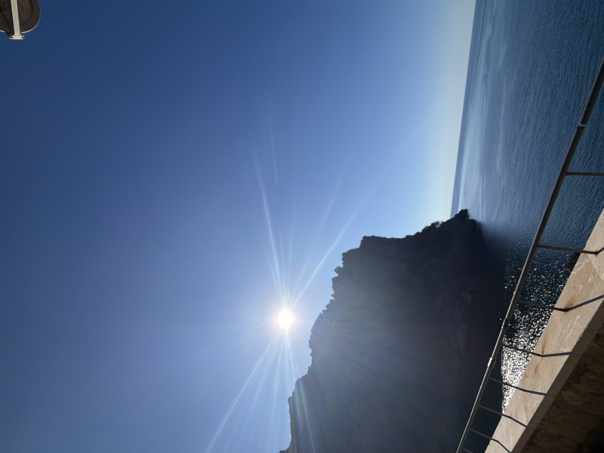 The image shows a coastal scene, likely taken on a sunny day, with the view angled upwards. A large, dark rocky outcrop extends into the water, with the sun positioned just behind it, creating a bright glare and radiating light beams. The sky is a clear blue, transitioning to a lighter hue near the horizon. A portion of the ocean is visible, appearing dark with slight reflections of the sunlight. A section of a railing or wall is present in the lower right corner, suggesting a viewpoint from a structure. Based on the location provided, the scene likely depicts a rugged stretch of coastline within the municipality of Escorca, Spain.