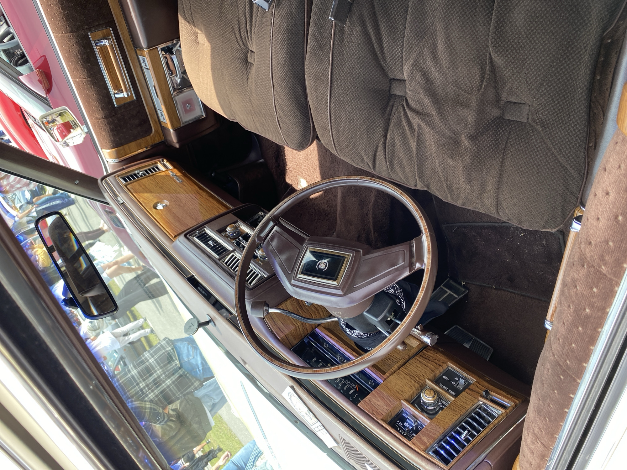 The image shows the interior of a vintage car, possibly a Cadillac, with plush brown velour seats and wood-grain trim. The dashboard features analog gauges and a large steering wheel. The exterior of the car is visible through the windshield and side windows, showing a crowd of people and other vehicles, suggesting an outdoor event like a car show or rally. The scene is bright and sunny, with a slight glare on the windshield. The car appears to be parked on an outdoor paved surface, possibly a street or parking lot. The presence of other vintage cars and a gathering of people indicates a public event. The setting appears to be an urban environment, potentially a city street or public square. Without additional context or landmarks, it is impossible to definitively place the image in Gdańsk, Poland.