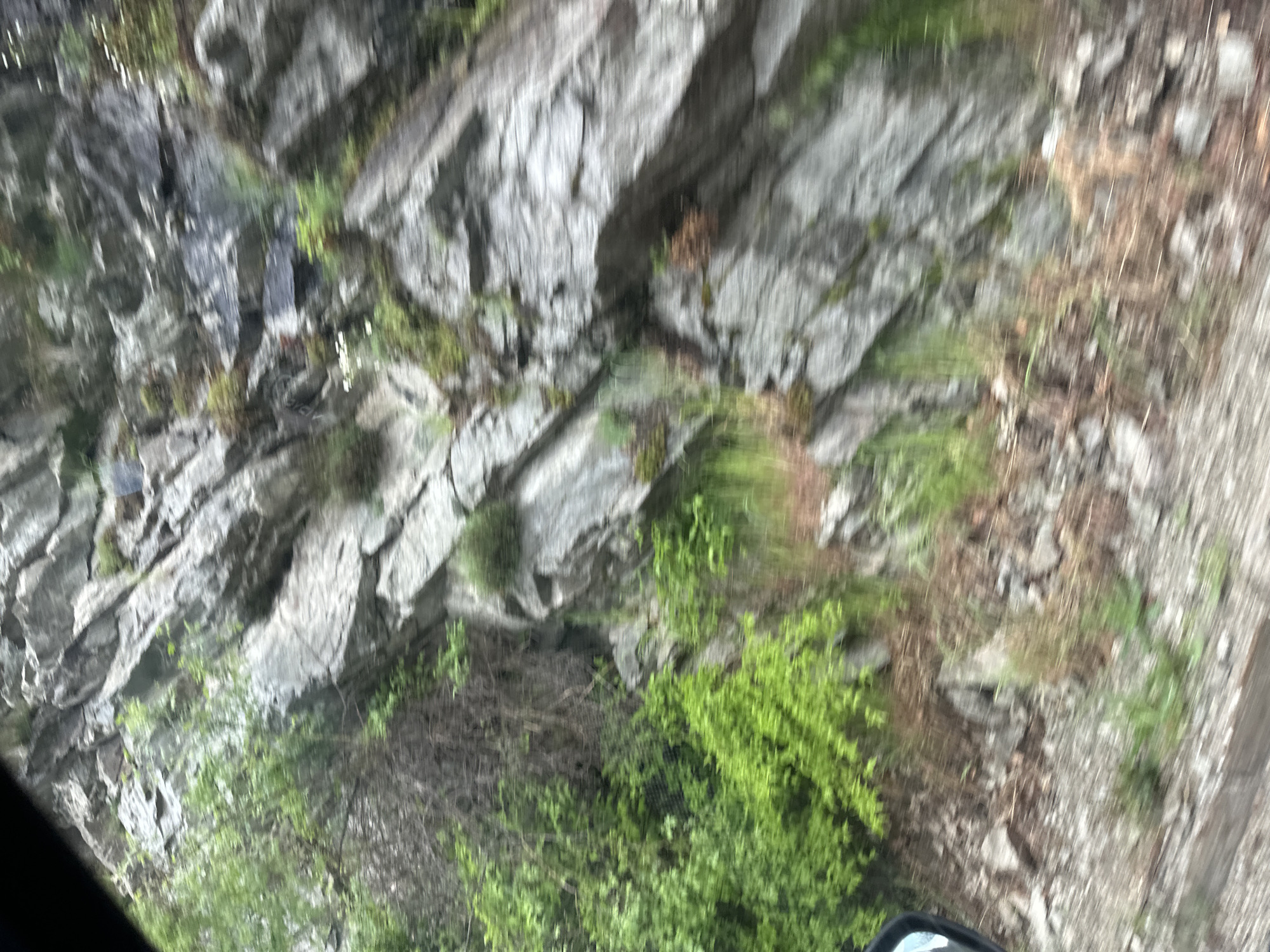 The image shows a rocky hillside with patches of green vegetation. The rock is gray and appears to be layered or fractured. Some small plants or moss are growing on the rock face. The ground at the base of the rock face is covered with vegetation, with some bare patches of soil or rock. The image appears to be taken from a moving vehicle. The location, Karnare, Bulgaria, is implied but not visually confirmed. No people are visible in the image.