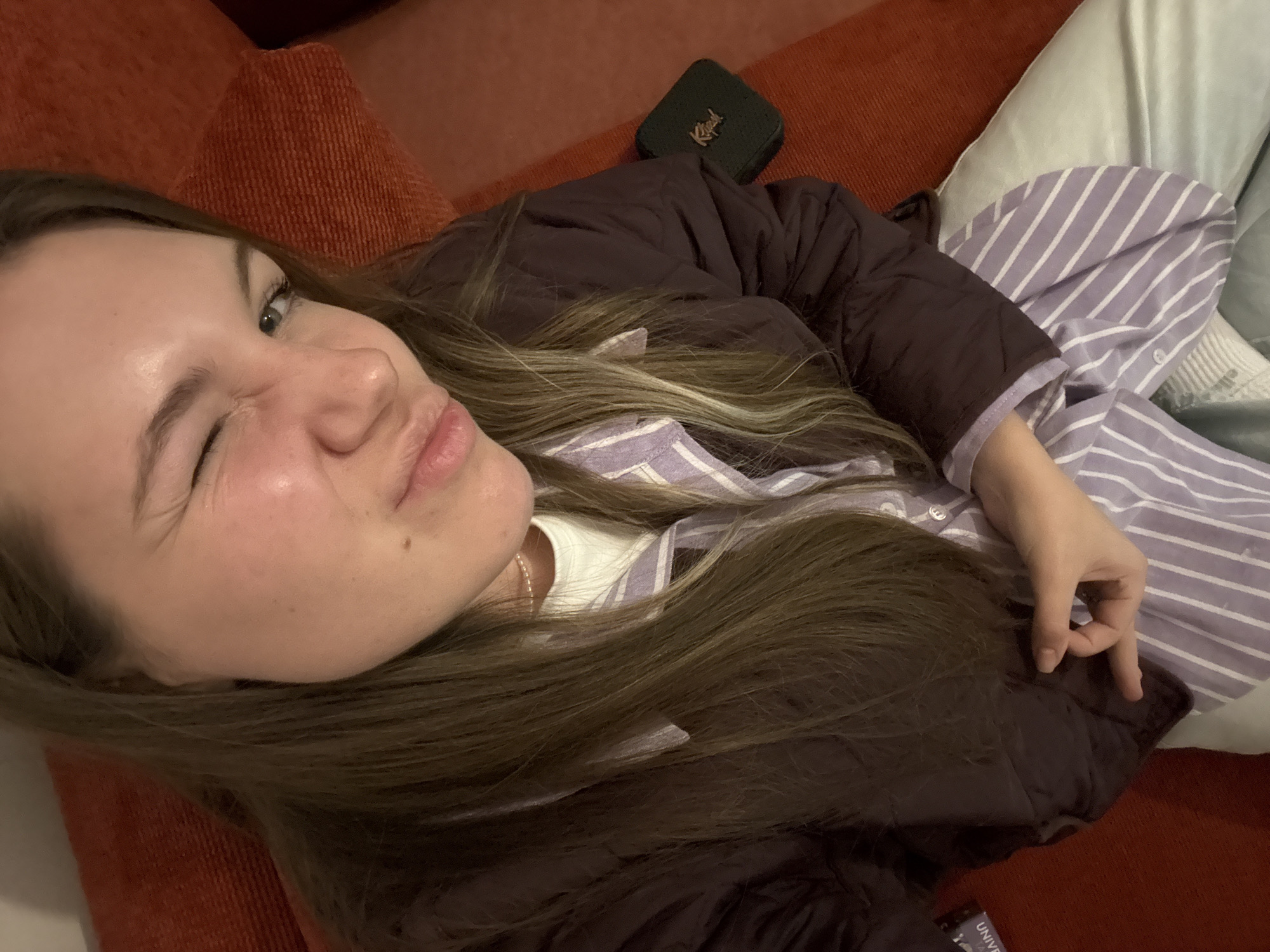 A young woman is captured in a close-up selfie, reclining indoors on a red or orange textured couch. She is winking playfully with her left eye and has her lips slightly pursed, conveying a casual and lighthearted expression. Her long, light brown hair is spread across the couch and her shoulders. She wears a dark brown or plum-colored quilted jacket over a light purple and white striped collared shirt, with a white t-shirt visible underneath. Light-colored pants are partially visible on her lower body. A thin silver-colored necklace is also visible around her neck.
Adjacent to her head, on the couch, rests a small, dark green rectangular bag or wallet featuring a stylized golden word, possibly a brand name such as "Kissell" or "Kassell". A light-colored pillow with subtle vertical stripes is also visible behind her. The indoor setting appears to be lit by artificial light. In the bottom right corner of the image, a tag is partially visible with the text "UNIVERSITY". The image reflects a relaxed moment. The scene is set in Vozdovac Urban Municipality, Serbia.