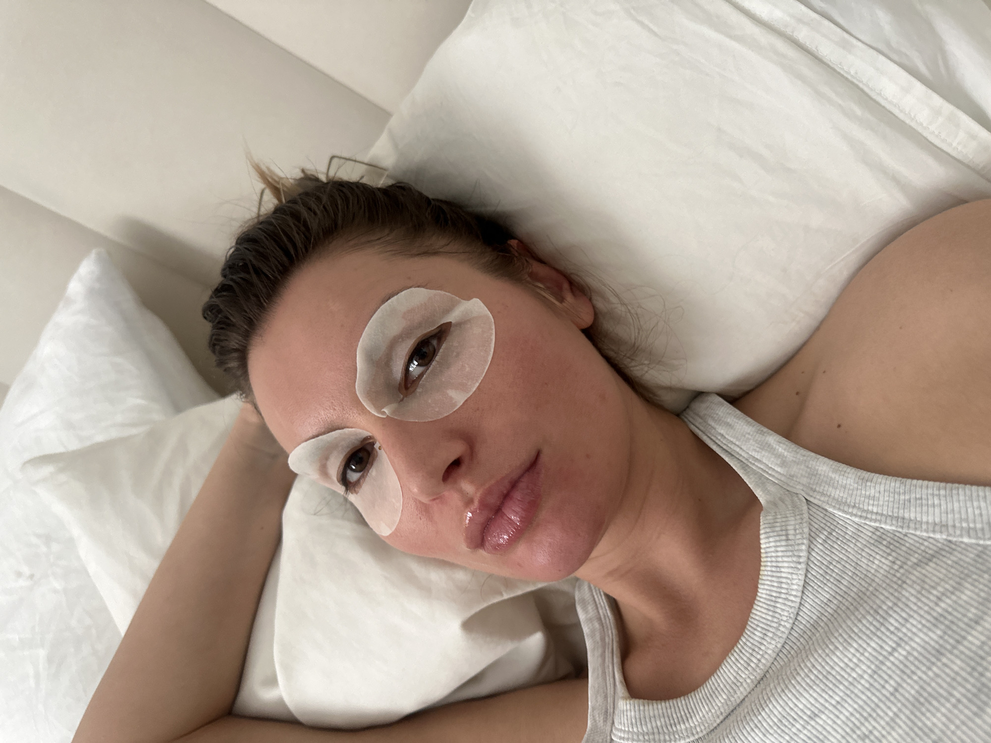 A close-up, high-angle image depicts a young woman lying comfortably on white bedding, likely in a bed indoors. She is looking directly at the camera with a calm and relaxed expression. Her light brown hair appears slightly disheveled or damp, pulled away from her face. She has two white, crescent-shaped eye patches applied under her eyes, indicating a skincare routine or beauty treatment. Her lips have a glossy appearance. She is wearing a light grey, ribbed tank top. The background is dominated by soft white pillows and sheets, contributing to a serene and comfortable atmosphere. The lighting is even and soft, suggesting an indoor setting. Her posture conveys a sense of rest or relaxation. The scene takes place in a private, domestic setting, possibly in Moscow, Russia. No visible text is present in the image.