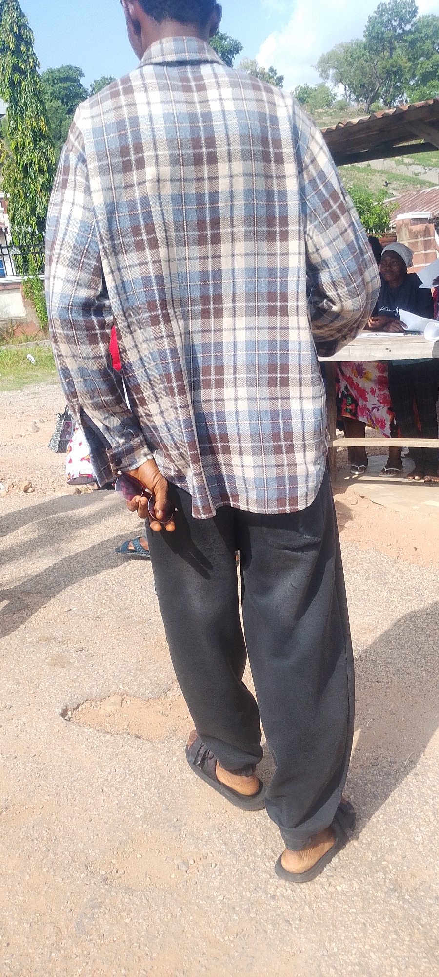 A man stands on a paved surface in Lokoja, Nigeria. He wears a plaid long-sleeved shirt, black sweatpants, and sandals. He holds sunglasses in his left hand. A woman sits at a table under a corrugated metal roof in the background. Other people are visible nearby. Green trees are in the background.