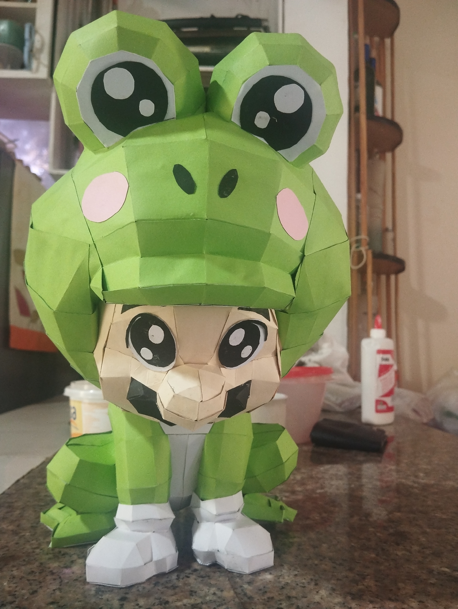 A papercraft figure of Mario in a frog suit sits on a countertop. The figure is made of polygonal paper pieces and depicts Mario with large eyes and a frog-shaped hood with bulging eyes and pink cheeks. The figure is green, white, tan, and black. In the background, a kitchen or dining area contains a cabinet, a shelving unit, a partially visible orange object, a container, a clear plastic container, glue, and other miscellaneous items. The location may be El Valle del Espíritu Santo, Venezuela.