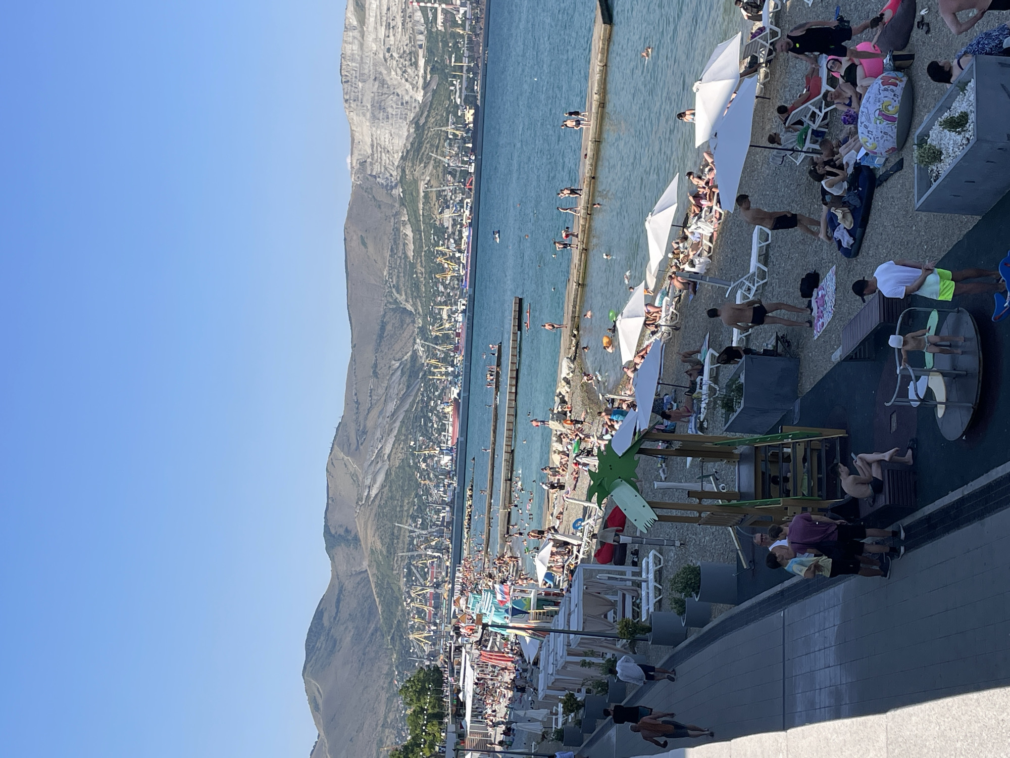 The image depicts a bustling beach scene under a clear blue sky, suggesting a warm, sunny day. A multitude of people are engaged in various beach activities. Some are sunbathing on lounge chairs, others are swimming or wading in the calm blue water, and many are walking along the pebble beach. Beach umbrellas provide shade for some of the sunbathers.
In the background, a large mountain range rises majestically, with a town or city built into its slopes, characterized by numerous buildings and industrial cranes, hinting at a port or industrial area. The sea is dotted with several small boats. The scene appears to be a popular tourist destination, given the density of people enjoying the recreational facilities. The overall atmosphere seems relaxed and cheerful, with people enjoying a day at the seaside. There is no visible text in the image. The rotation of the image suggests it was taken vertically.