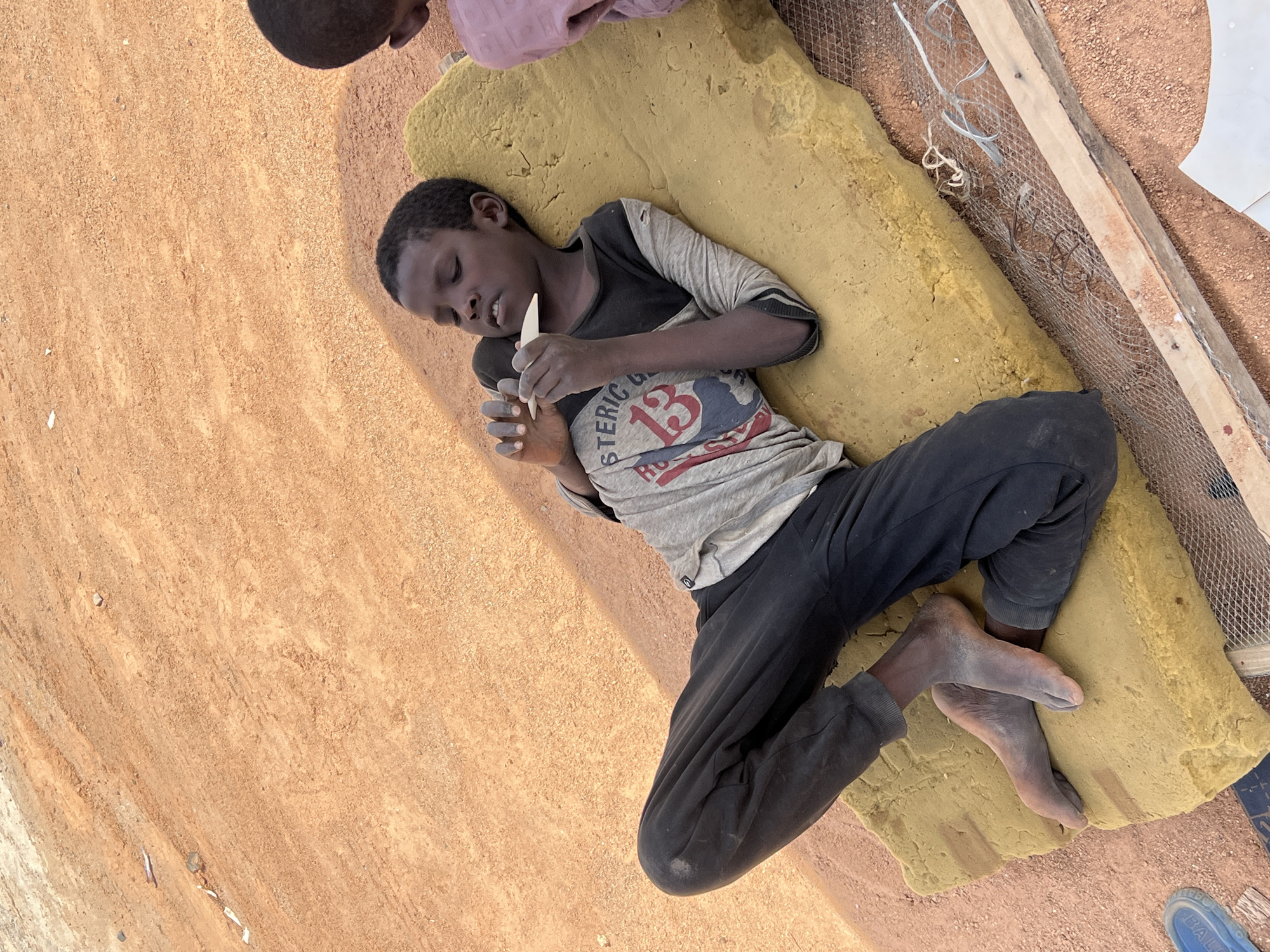 A young boy lies on a yellow foam mattress placed on a dirt ground in Barakin Lerawi, Nigeria. He is wearing a grey T-shirt with the text "STERIC" and the number "13" printed on it, and black sweatpants. He holds a small, light-colored object in both hands. Another person is partially visible in the upper left corner. Next to the mattress are wooden boards and wire mesh.