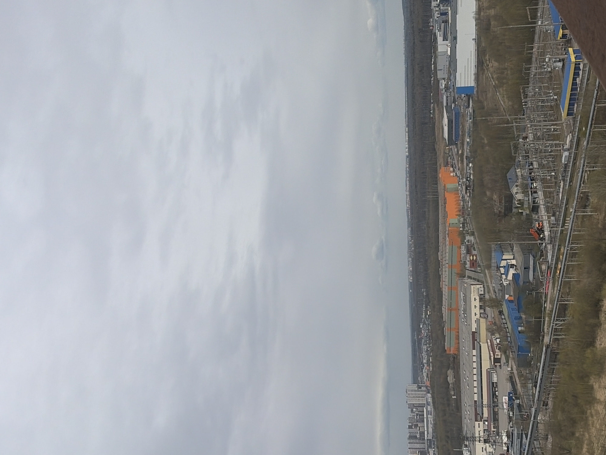 The image presents a wide, elevated view of an industrial or commercial landscape under an overcast sky. The upper two-thirds of the frame is dominated by a vast expanse of light gray, cloudy sky, suggesting a diffused and muted light, indicative of an overcast day. A distant, hazy horizon line is visible beneath the clouds, showing a cityscape stretching across the far distance.
The lower third of the image captures a functional urban periphery. This area is characterized by a series of large, rectangular buildings, consistent with warehouses or factories. Some buildings feature prominent orange roofs, while others are white or blue. A significant electrical substation or industrial complex is visible, replete with numerous pylons, transformers, and intricate electrical infrastructure. Interspersed among the buildings and utility corridors are patches of sparse vegetation and bare trees, hinting at early spring or late autumn foliage. A network of roads or pathways connects the various structures. In the far bottom right, a blurry brown element suggests part of the structure from which the photograph was taken.
No people are visible, and no specific event or activity is discernable; the scene is static. The overall impression is one of an expansive, utilitarian landscape. Faint Cyrillic text, possibly "КОРПУС" (KORPUS), is barely visible on one of the large buildings. Given the location context of Saint Petersburg, Russia, the industrial development and architectural styles are consistent with a Russian urban environment. The time of day appears to be daytime, with no strong shadows indicating a direct sun source.