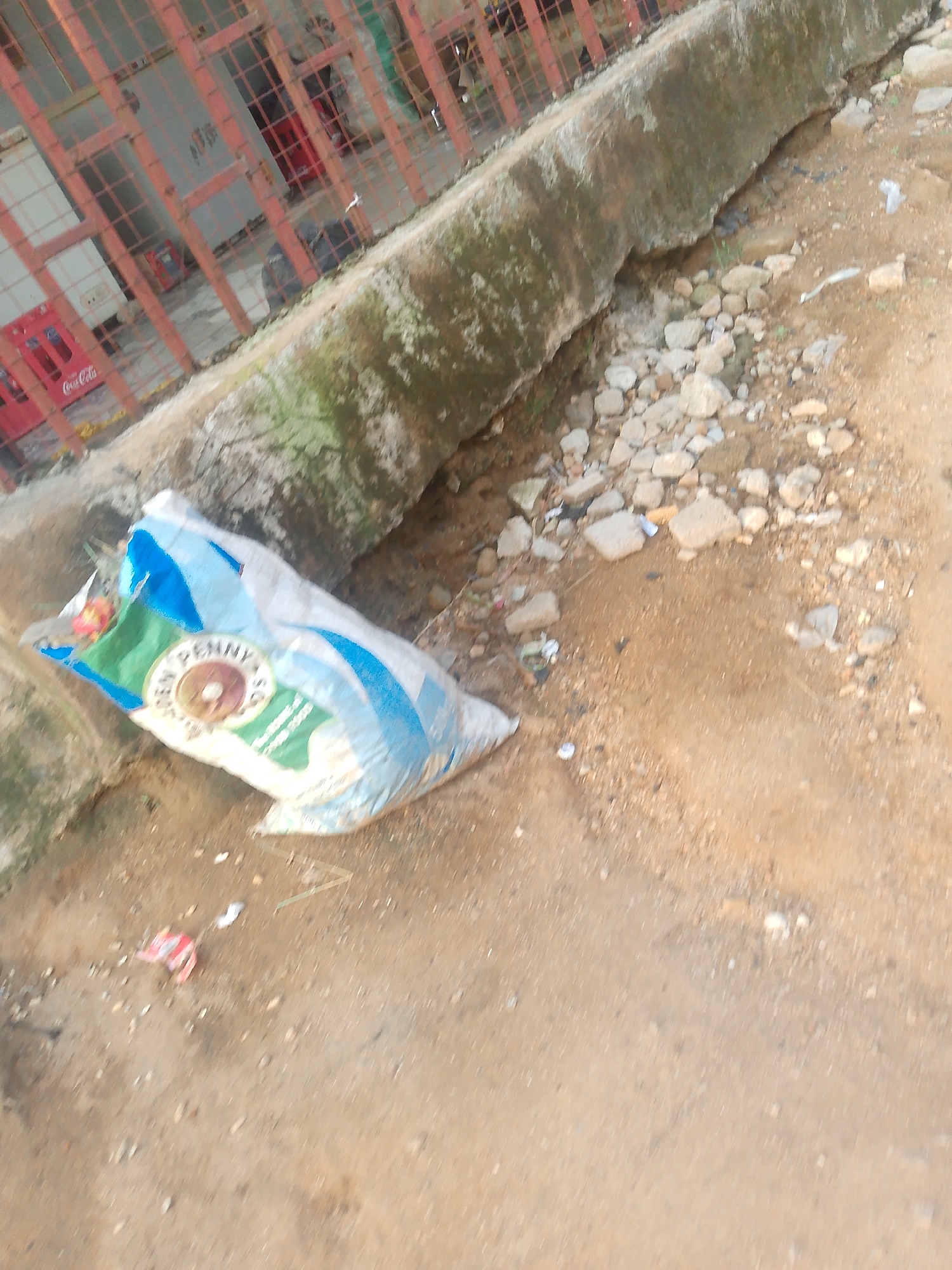 In the city of Jalingo, Nigeria, a discarded white and blue plastic bag lies on a dirt path next to a concrete barrier. The bag appears to contain loose material, possibly grain, and has some branding visible on its surface, including the words "DEN PENNY SG." Scattered around the bag are small stones and debris, suggesting an outdoor, possibly unkempt, location. Behind the barrier, a red metal fence encloses an area with what appears to be a structure or building, and red Coca-Cola crates are visible through the fence. The ground is a mix of sand and small pebbles.