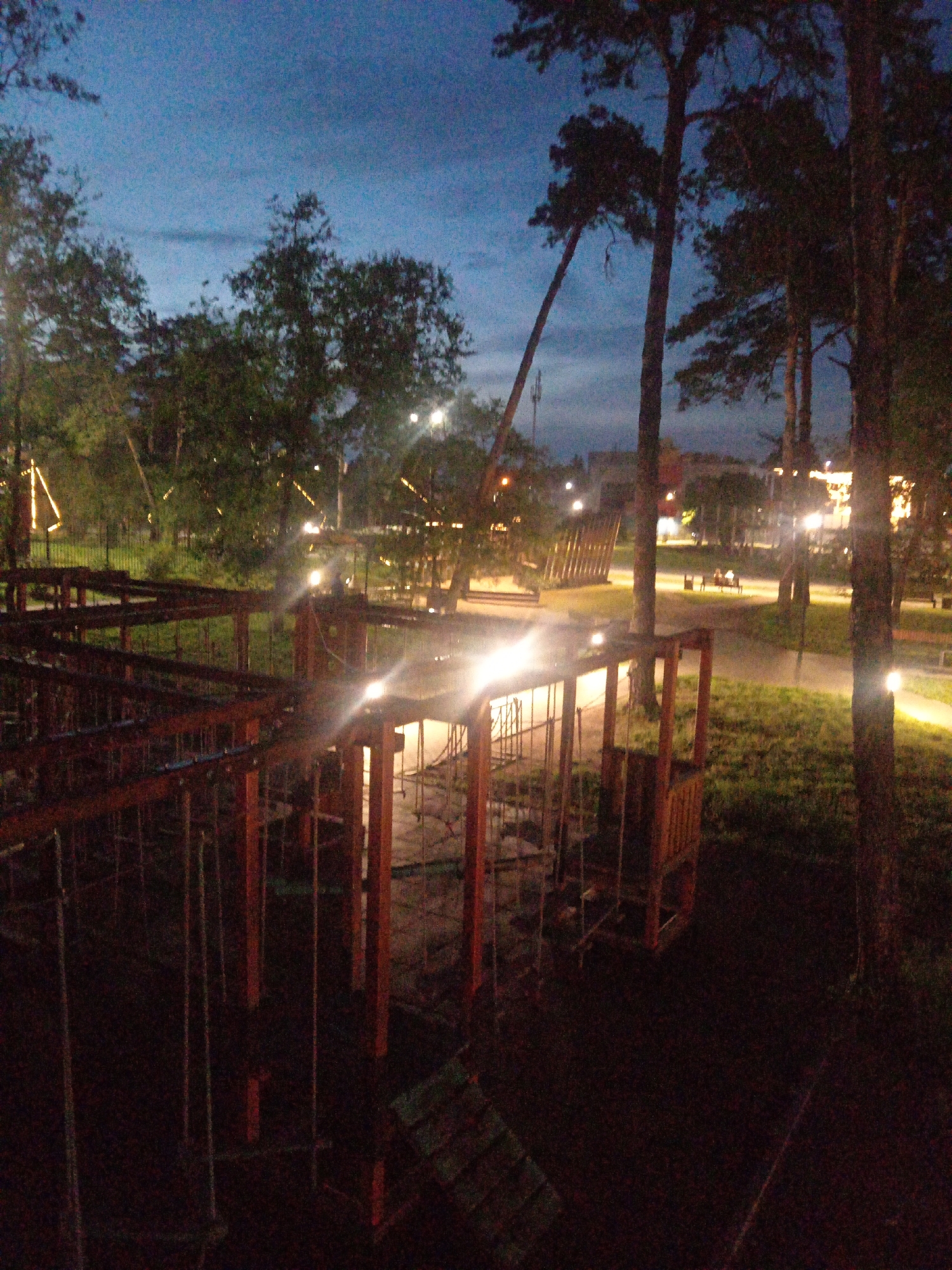 The image depicts an outdoor adventure park or obstacle course at dusk in Belogorsk, Russia. The sky is a deep blue, transitioning to lighter hues near the horizon, with a few scattered clouds. Tall pine trees are prominent in the foreground and midground, their dark silhouettes contrasting with the twilight sky.
In the foreground, a wooden structure, likely part of the obstacle course, is visible. It features a wooden frame with ropes and netting, suggesting it's a climbing or traversing element. The structure is dimly lit, with some lights from within and around it creating bright flares.
Further back, illuminated pathways wind through grassy areas and among trees. Several lampposts are visible along these paths, casting pools of light. In the midground, a couple of people are seated on a bench, their figures indistinct due to the low light conditions and distance. Other structures and trees are scattered throughout the park.
In the background, buildings with illuminated windows suggest the proximity of a residential or commercial area. The overall atmosphere is serene and somewhat mystical, with the artificial lights of the park juxtaposed against the natural beauty of the trees and the fading daylight.