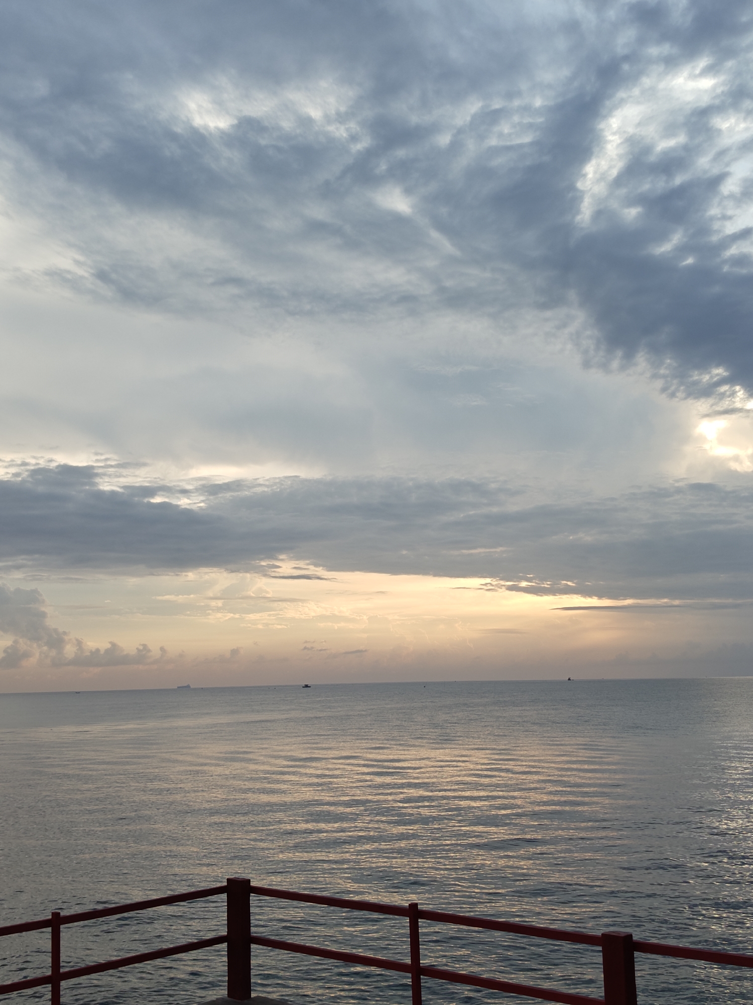 The image captures a serene seascape in Lhokseumawe, Indonesia, likely at dawn or dusk. The sky is a canvas of soft hues, with layers of gray and white clouds gradually giving way to a warm, peachy glow near the horizon, suggesting the sun is either rising or setting. Sunlight peeks through the clouds, casting gentle reflections on the calm water. The sea itself appears tranquil, with subtle ripples creating a textured surface.
In the distance, several small boats or ships are visible on the horizon, indicating maritime activity. The foreground features a red railing, part of a pier or walkway, providing a sense of perspective and framing the expansive view of the ocean. The overall atmosphere is peaceful and contemplative.