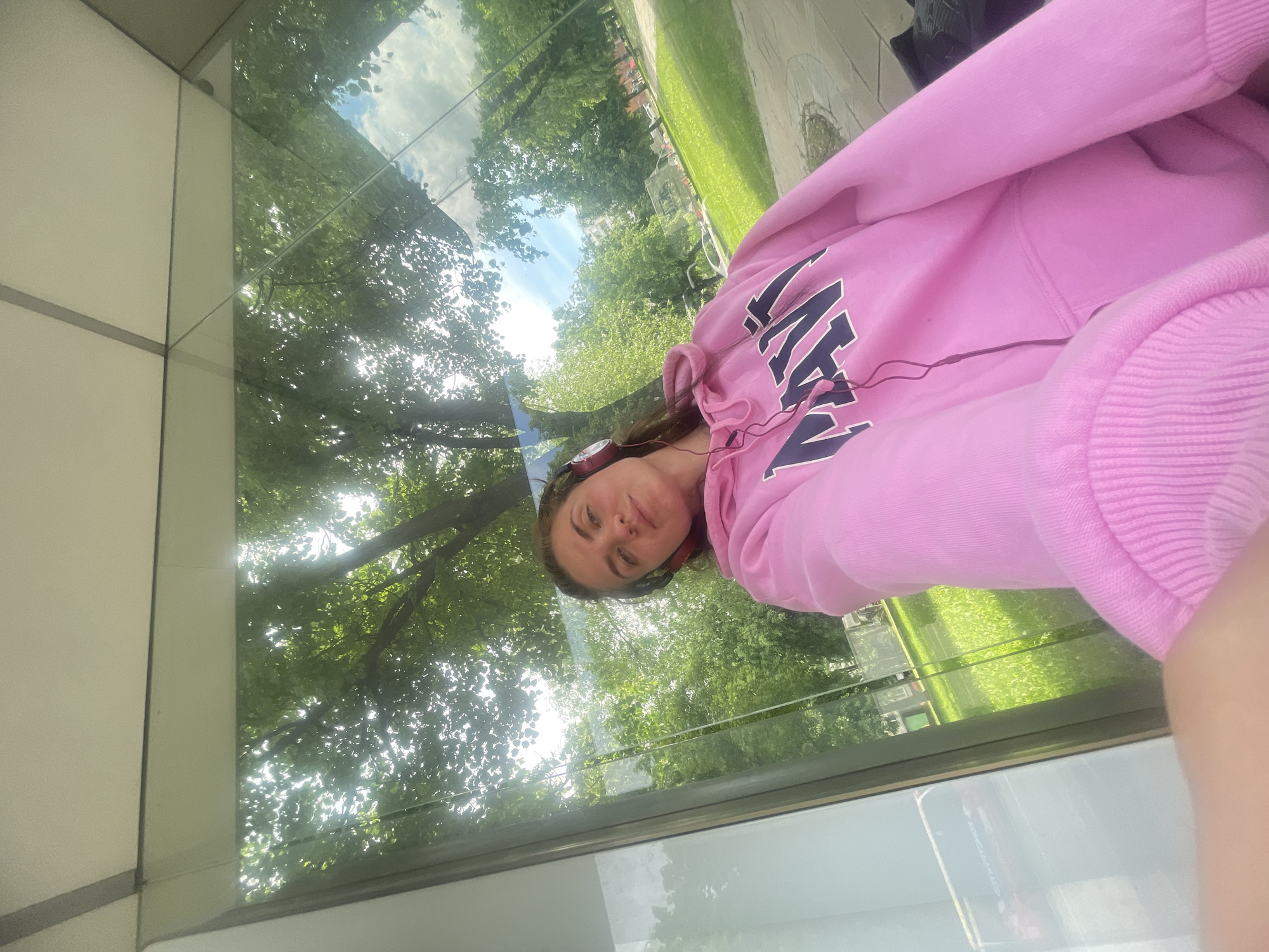 A woman is wearing a pink hoodie and headphones while standing in front of a window reflecting trees and sky. The location appears to be Moscow, Russia.