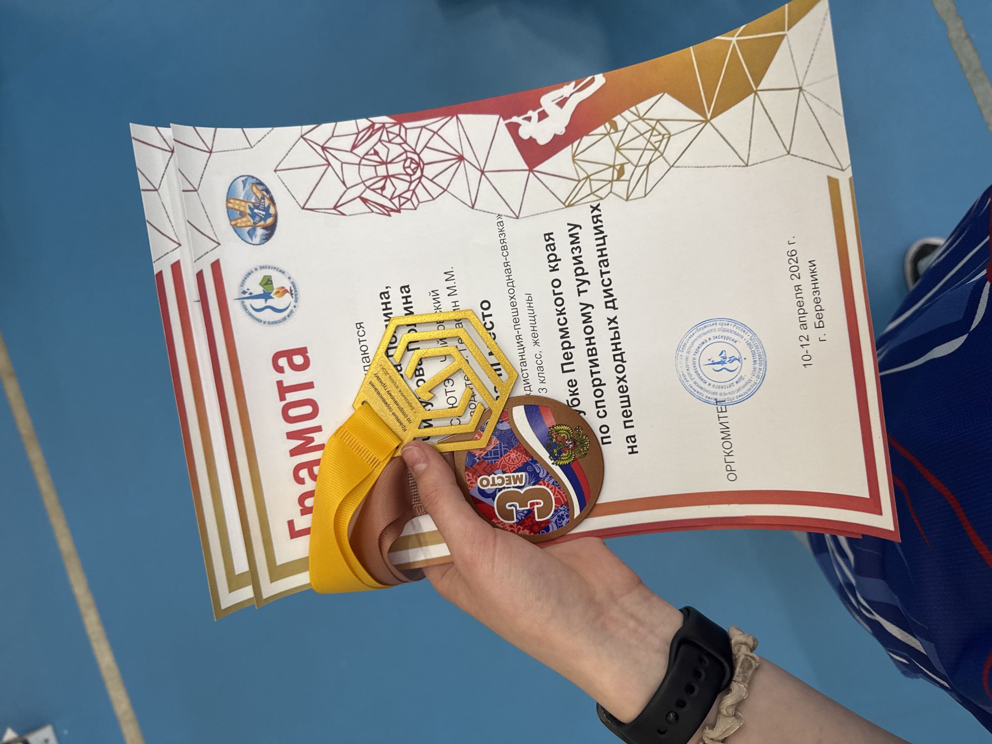 The image shows a person holding a certificate and a medal. The certificate is titled "Грамота" and appears to be an award for sports tourism in the Perm region of Russia, specifically for pedestrian distances. It dates to April 10-12, 2026, in Berezhniki. The medal is for 3rd place. The person is wearing a black smartwatch. They are holding the certificate in their left hand. The location cannot be determined from the image, although the certificate indicates it is related to the Perm region of Russia, and possibly the city of Berezhniki. There is no indication of the city of Usolye.