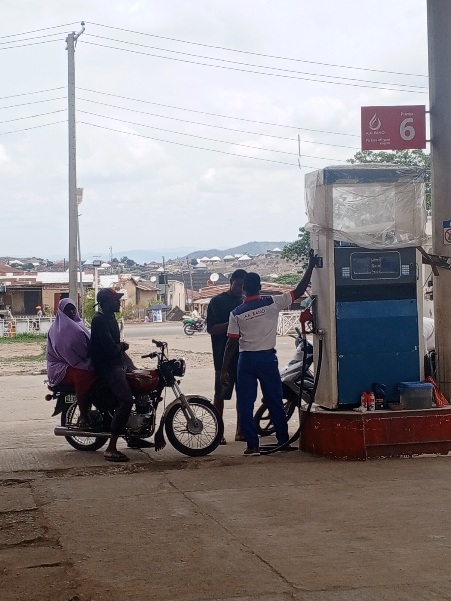 At a gas station in Unknown, Unknown, a man wearing a white and blue uniform is operating a gas pump, which is labeled "Pump 6" and covered in plastic. He is facing the pump and appears to be holding the nozzle. To the left of the pump, there are two men and a woman sitting on a red motorcycle. The woman is wearing a purple headscarf. Another man is standing next to the motorcycle, facing the pump. Behind the gas station, there is a view of buildings and hills. In the foreground, the ground is paved and uneven. A tall utility pole with power lines stands to the left of the gas station. Various containers and bottles are placed on the base of the gas pump.