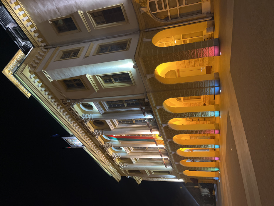 The image depicts the Municipality of Sliven building in Sliven, Bulgaria, at night. The multi-story classical-style building features a light-colored facade, illuminated by a combination of warm yellow and white lights. The upper level is adorned with a series of columns with Corinthian capitals, between which tall windows are visible. A frieze above the columns displays the text "ОБЩИНА СЛИВЕН" and "MUNICIPALITY OF SLIVEN." On the roofline, two flags are present: the Bulgarian flag (white, green, red vertical stripes) and the European Union flag (blue with yellow stars). A large, vertically-hung Bulgarian flag is prominently displayed from a central balcony on the upper level. The lower level of the building features a series of robust arches, forming a covered walkway, which are lit from below with warm yellow light. The bases of several columns supporting these arches are individually illuminated by distinct colored spotlights, including pink, blue, green, and red. The ground in front of the building is a paved plaza, exhibiting geometric patterns, and is free of people or vehicles. A streetlight is partially visible on the far left. The sky is dark, indicating nighttime conditions.