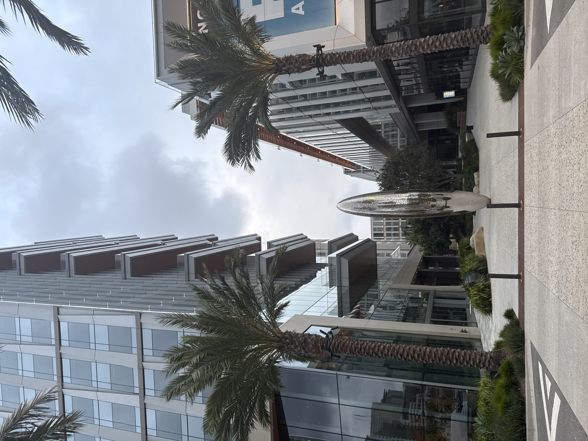 The image depicts a modern architectural scene, likely in a commercial or business district, possibly in San Diego, United States. The view is angled upwards, focusing on two tall buildings with contemporary designs.
The building on the left features multiple levels of reflective glass windows and protruding architectural elements, creating a stepped or layered effect. The building on the right is taller and appears to have a more streamlined design, with repeating horizontal lines. Palm trees are strategically placed in the foreground, partially obscuring the buildings and adding a tropical aesthetic to the scene.
In the center of the frame, a large, shiny, elliptical sculpture sits on a paved area. To the right of the sculpture, there are manicured plants along the edge of the pavement. The sky is overcast, suggesting a cloudy day. The environment is clean and well-maintained, suggesting a high-end or upscale area. There are no visible people or vehicles.