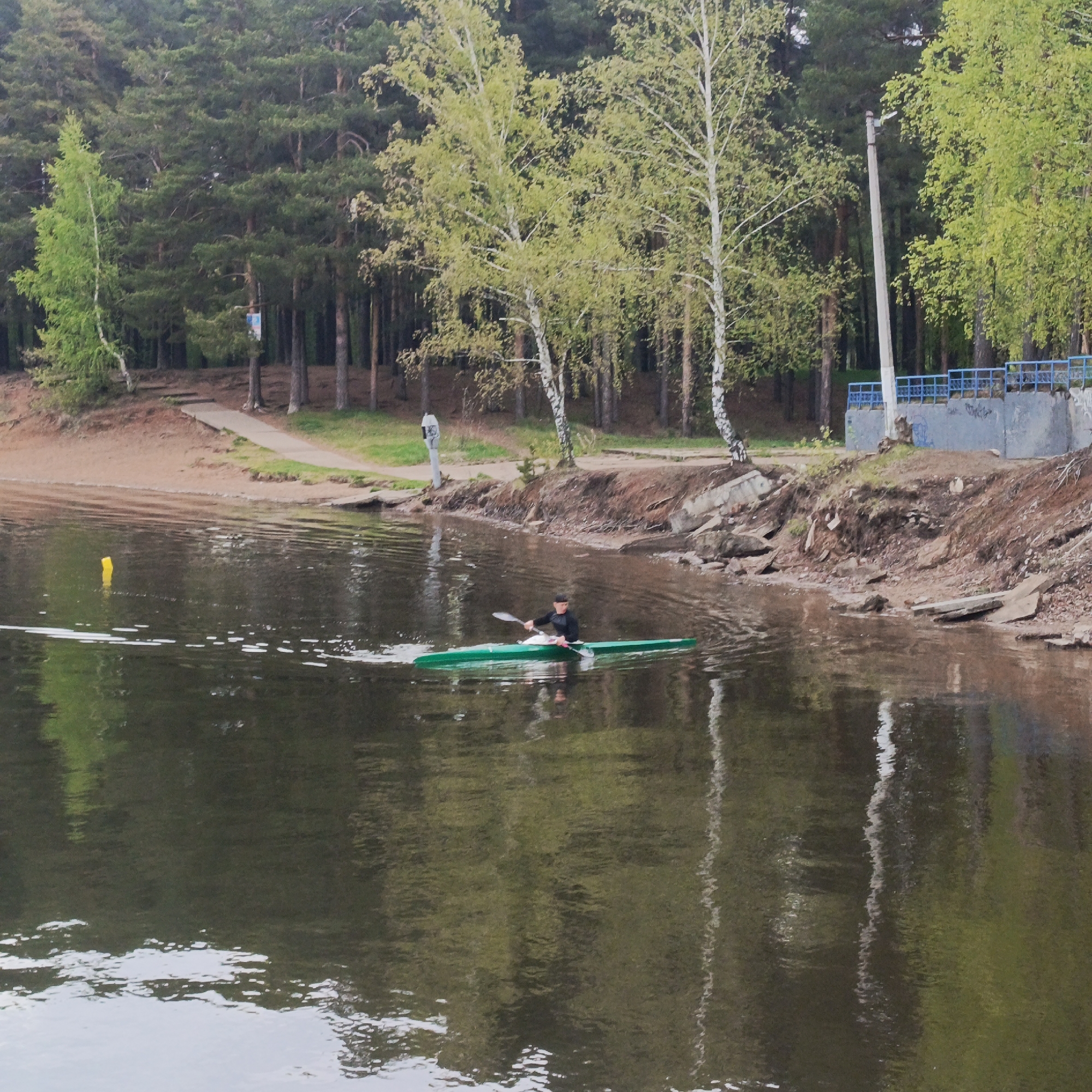 The image depicts a person kayaking on a body of water, likely a lake or large pond. The kayaker is positioned in the center of the frame, paddling a green kayak.
The surrounding environment includes a shoreline with trees. Pine trees are visible in the background. The image features a pier, and a concrete retaining wall. There is a yellow buoy in the water to the left of the kayaker.
The scene appears to be at the city of Chaikovsky, Russia.