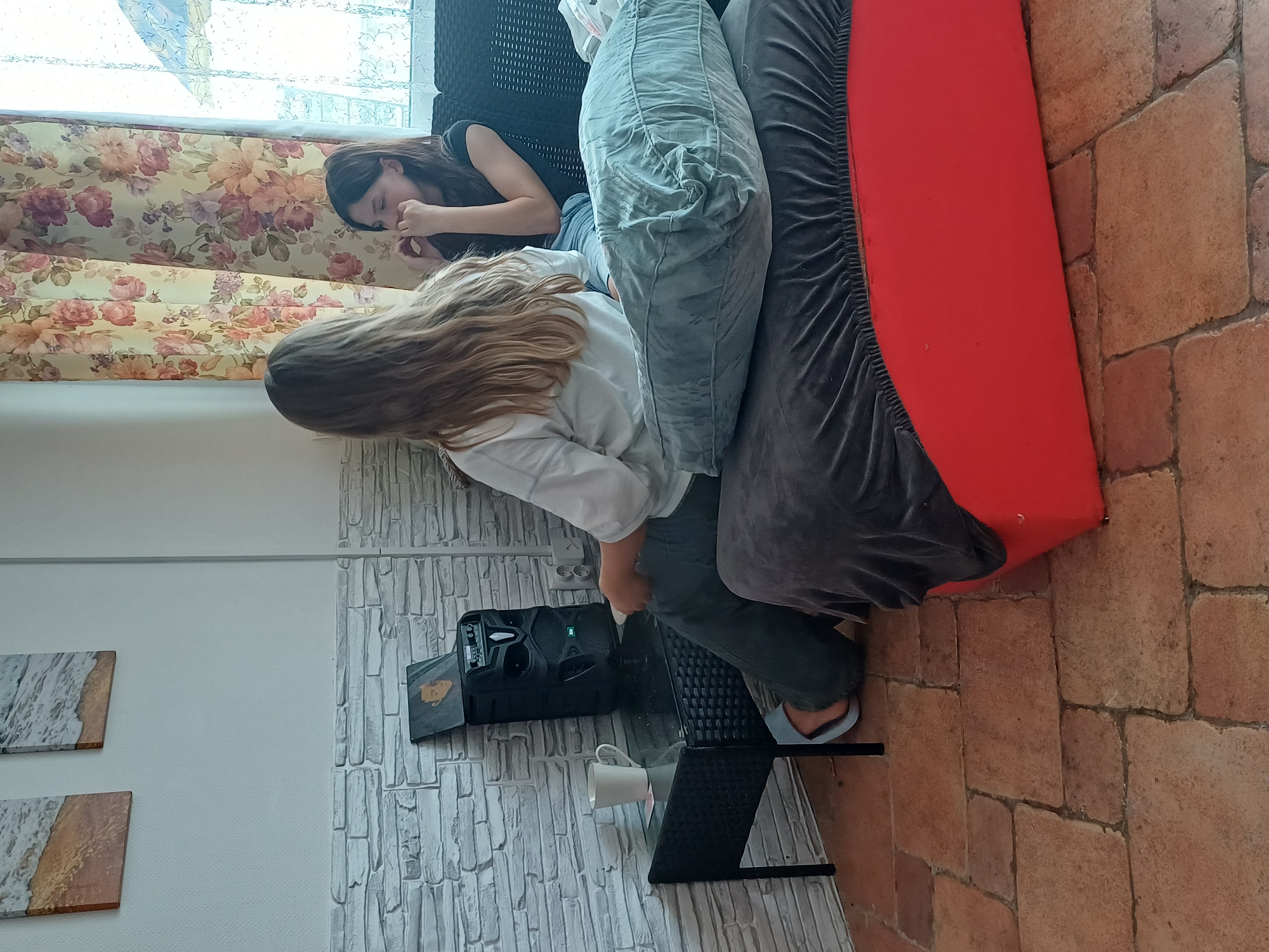 The image shows two young women lounging on a sofa in a brightly lit room. The woman on the left, with dark hair, is lying down with her head on a pillow, appearing to be sleeping or resting her eyes. She is wearing a black t-shirt. The woman on the right, with long blonde hair, is sitting up and leaning forward. She is wearing a white shirt and dark pants. She is looking down, possibly at a device or object on the coffee table in front of her.
In front of the sofa is a black coffee table with a glass top. On the table sits a white mug and a black electronic device, possibly a portable speaker or a projector, with its lid open. The room has large windows with floral curtains, suggesting a daytime scene. The floor is tiled with terracotta-colored bricks. In the background, there are decorative wall panels with abstract patterns of what appears to be marble and sand. The overall impression is one of a relaxed, casual atmosphere. The lighting suggests it is daytime, and there are no indications of specific weather conditions. The setting appears to be a home or a living space, possibly a vacation rental given the tiled flooring and decorative panels.