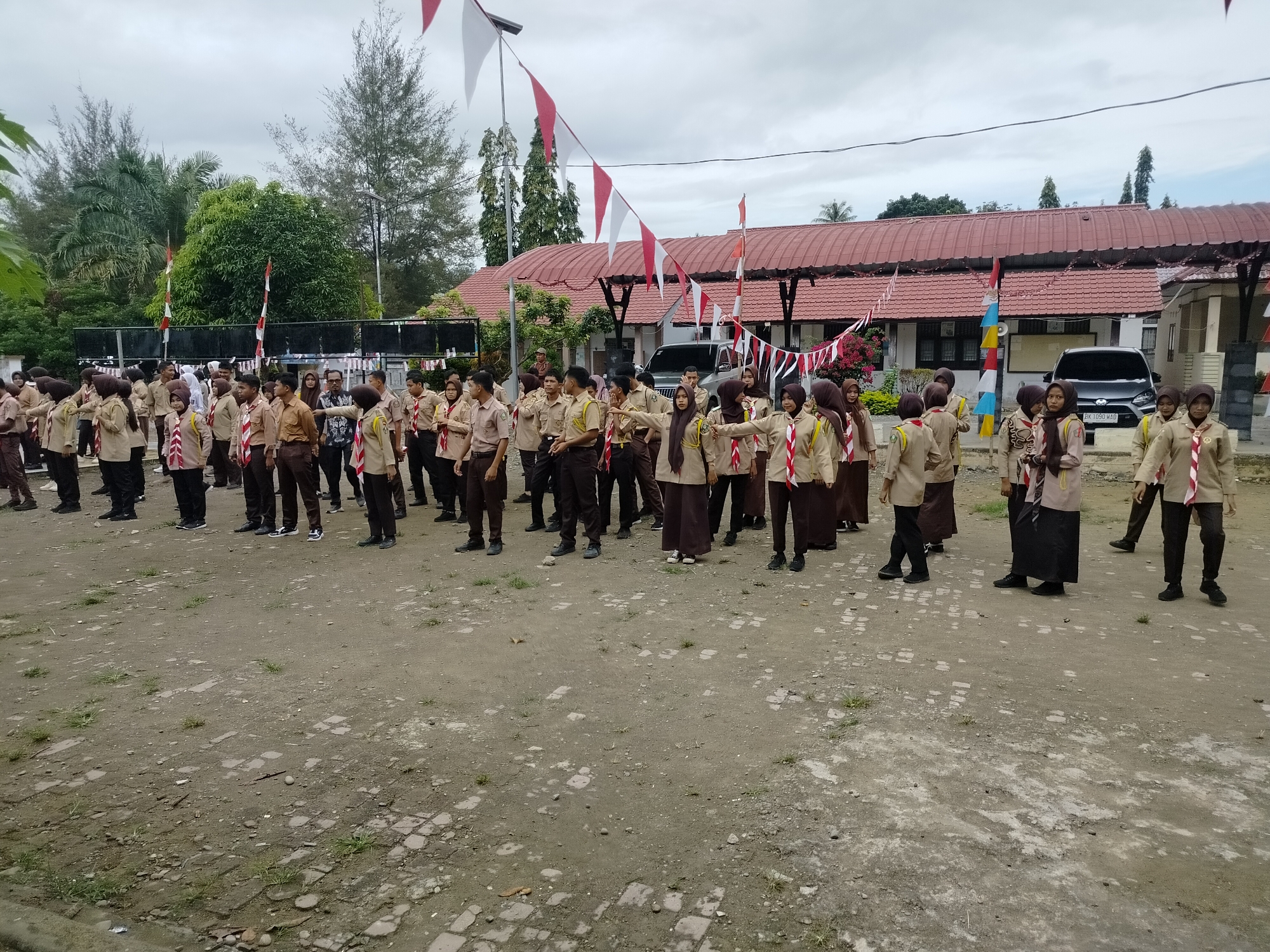 This is a photograph of a group of people, likely students, dressed in scout uniforms standing in an outdoor area. The uniforms consist of tan shirts, brown pants or skirts, and red and white neckerchiefs. Many of the individuals are wearing headscarves, indicating a likely Islamic context. In the background, there is a building with a red tiled roof and a covered walkway. Flags, predominantly red and white, are strung across the area, suggesting a festive or ceremonial occasion. Two cars are parked in the background. The ground is a mix of dirt, gravel, and some patches of grass and small puddles. The scene appears to be in a school or community setting. The presence of Indonesian flags suggests the location is in Indonesia, and potentially the city of Purwodadi.