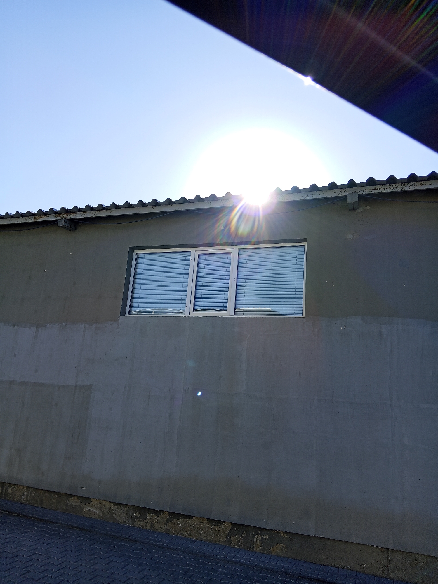 The image shows a close-up view of a building's exterior wall with a window, under a bright sky. The building has a flat, gray, concrete facade. A horizontal band of a different texture, appearing rougher and lighter gray, runs along the bottom of the wall. Above this band, the wall is smoother and darker gray. There is a window with white trim and horizontal blinds installed in the upper portion of the wall. A corrugated roofline is visible above the window. The sun is shining intensely, creating lens flare and a bright glare in the sky, partially obscured by an angled dark object in the upper right corner of the image. The ground in the foreground appears to be paved with a dark, patterned material. The scene is devoid of people or any immediate action. The overall environment suggests an outdoor, possibly urban, setting, though no specific city landmarks are identifiable in the image itself.