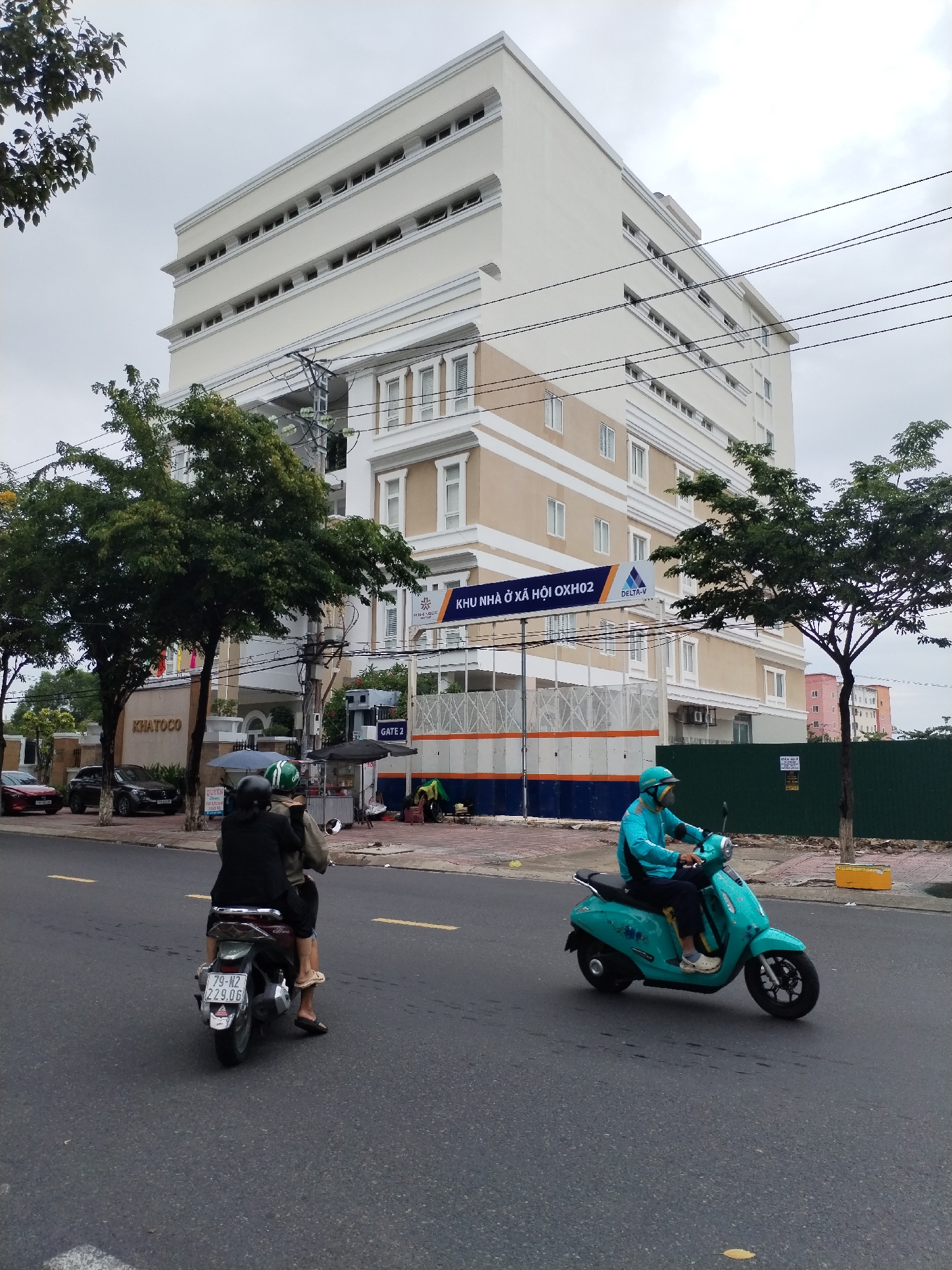 The photograph captures a street scene in Nam Nha Trang Ward, Vietnam, featuring a large multi-story building dominating the background. The building is predominantly white, with horizontal beige bands separating sections of windows, and is labeled "KHU NHÀ Ở XÃ HỘI OXH02 DELTA-V" on its lower portion. In front of the building, construction is underway, suggested by the blue and orange barriers.
Two motorbikes are the focal point in the foreground. The first, further away, carries two individuals: the driver wears a dark jacket and the passenger is dressed in lighter colors. The second motorbike, closer to the camera, is driven by a person wearing a turquoise jacket and helmet.
The street is grey asphalt with yellow lane markings. On the left side, several parked cars are visible in front of the building entrance, which bears the inscription "KHATOCO" above the entrance. Green trees frame the building on both sides. Overhanging electrical wires stretch across the sky. The weather appears overcast.