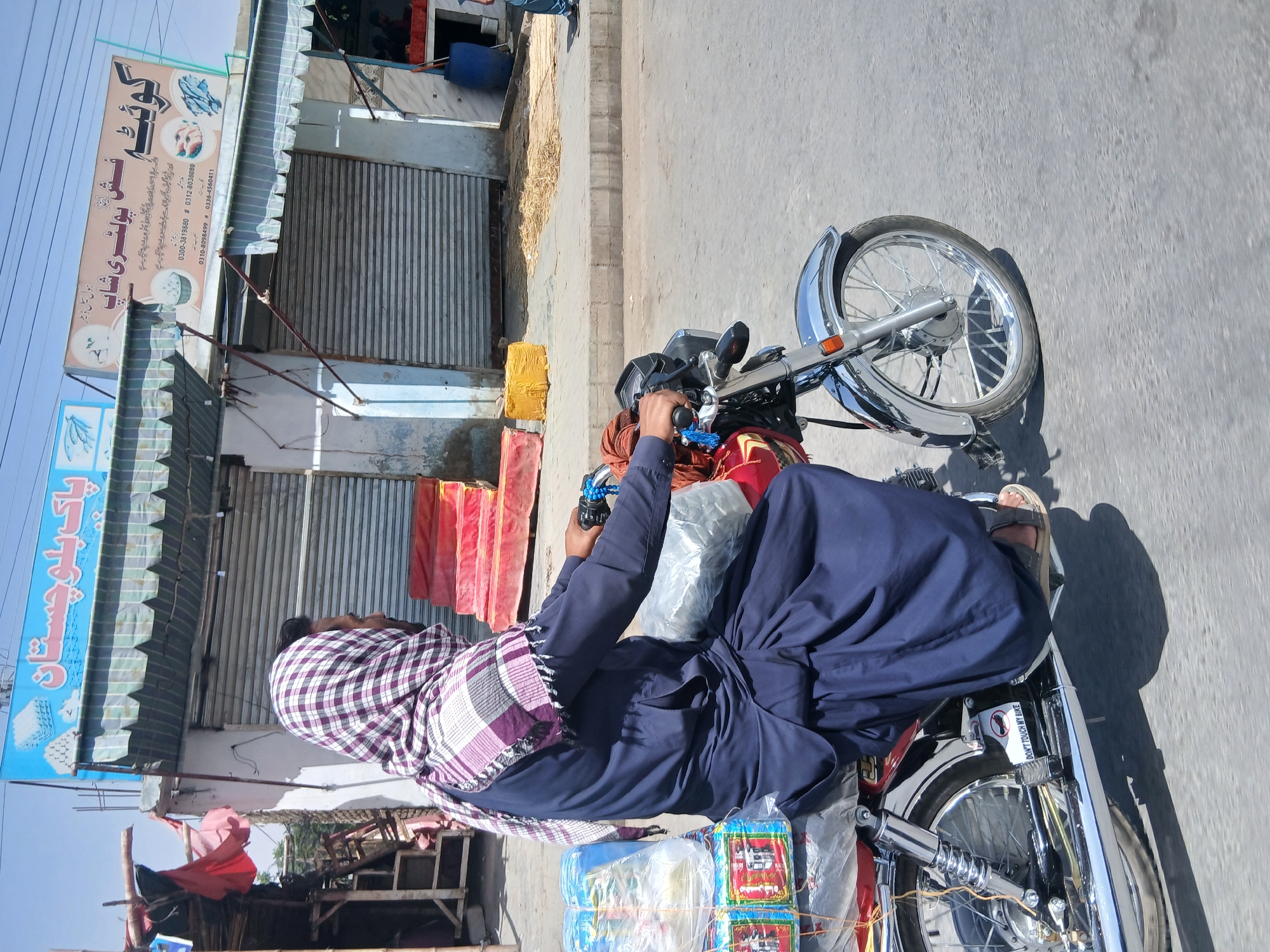 The image shows a man riding a motorcycle on a street in Quetta City Tehsil, Pakistan. The man is wearing a blue robe and a checkered headscarf. He is carrying packages of goods on the motorcycle. Behind him, there is a shop with a sign that reads "کوئٹہ فش پولٹری شاپ" (Quetta Fish and Poultry Shop). The shop has its shutters down.