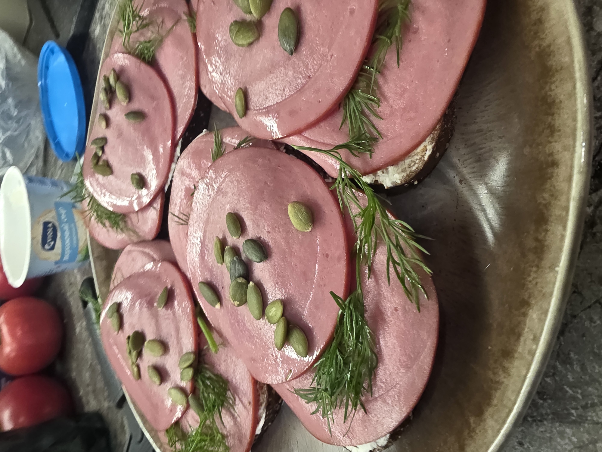 A close-up, overhead shot captures a plate of open-faced sandwiches, likely prepared for a meal or snack. Each sandwich consists of a base of dark bread, generously topped with a creamy spread, layered with several thin slices of pink, cured meat, and garnished with fresh dill sprigs and scattered pumpkin seeds. The sandwiches are arranged on a textured, grey-brown plate with a light-colored rim. In the blurred background, there are red tomatoes, a white plastic cup with a blue lid, and a blue plastic lid. The overall scene suggests a casual, home-cooked meal setting. The location is not specified in the image.