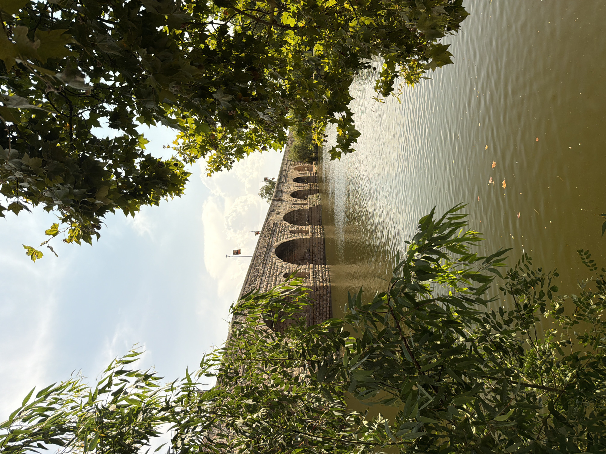 The image shows a view of the Roman Bridge of Mérida, Spain, with lush green trees framing the foreground. The stone bridge, with its multiple arches, spans across a calm body of water, reflecting the sky and surrounding greenery. Two flags are visible atop the bridge. The sky is partly cloudy with scattered white clouds. The water surface is rippled, with a few fallen leaves scattered on it. The scene is bathed in natural sunlight, suggesting it was taken during the daytime. The overall impression is one of natural beauty and historical grandeur.
