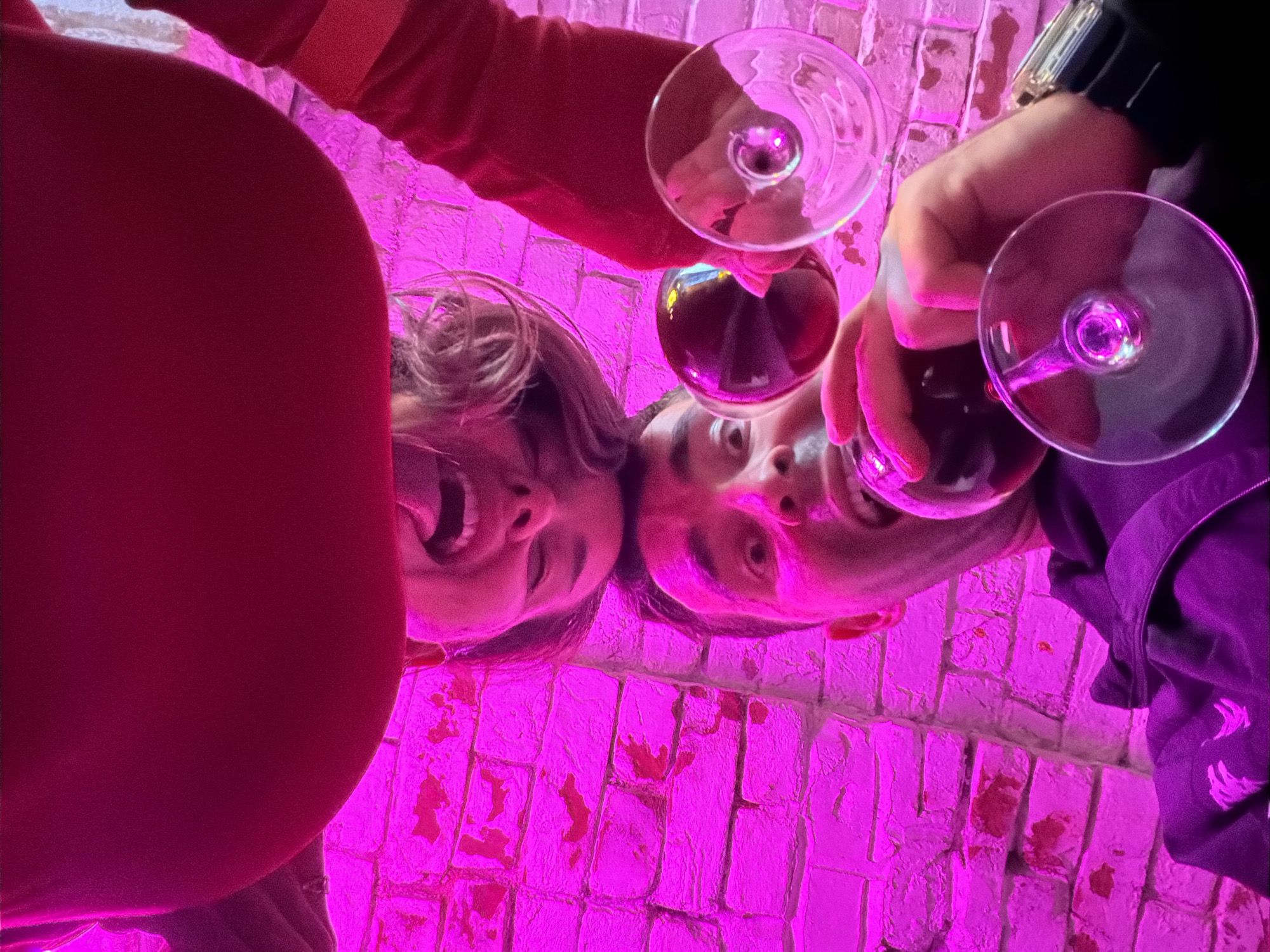 The image shows two people holding wine glasses, possibly toasting. A woman on the left side of the frame is laughing with her mouth open. A man is on the right side of the frame. The scene is lit with a bright, pink or purple light. The background appears to be a brick wall also illuminated with pink lighting. The location in the city of Krasnoyarsk, Russia is not evident from the image.