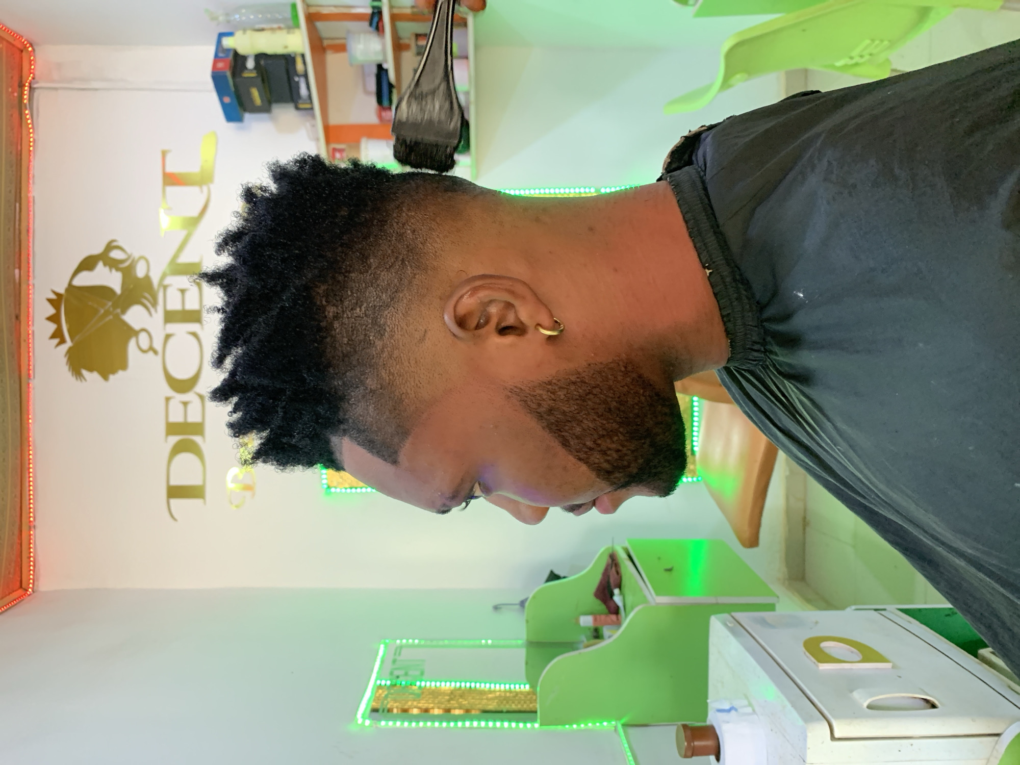 A person is in a barber shop, receiving a hair coloring treatment. The shop appears to be located in Ikorodu, Nigeria, judging from the "DECENT GENT" salon logo. A black hair dye applicator brush is hovering near the person's head. The person has a short, textured hairstyle and a beard. They are wearing a black garment or cape to protect their clothes from the dye. The shop is brightly lit with LED lighting. There are shelves with barbering products and green stools or chairs. A workstation with green accents is visible.