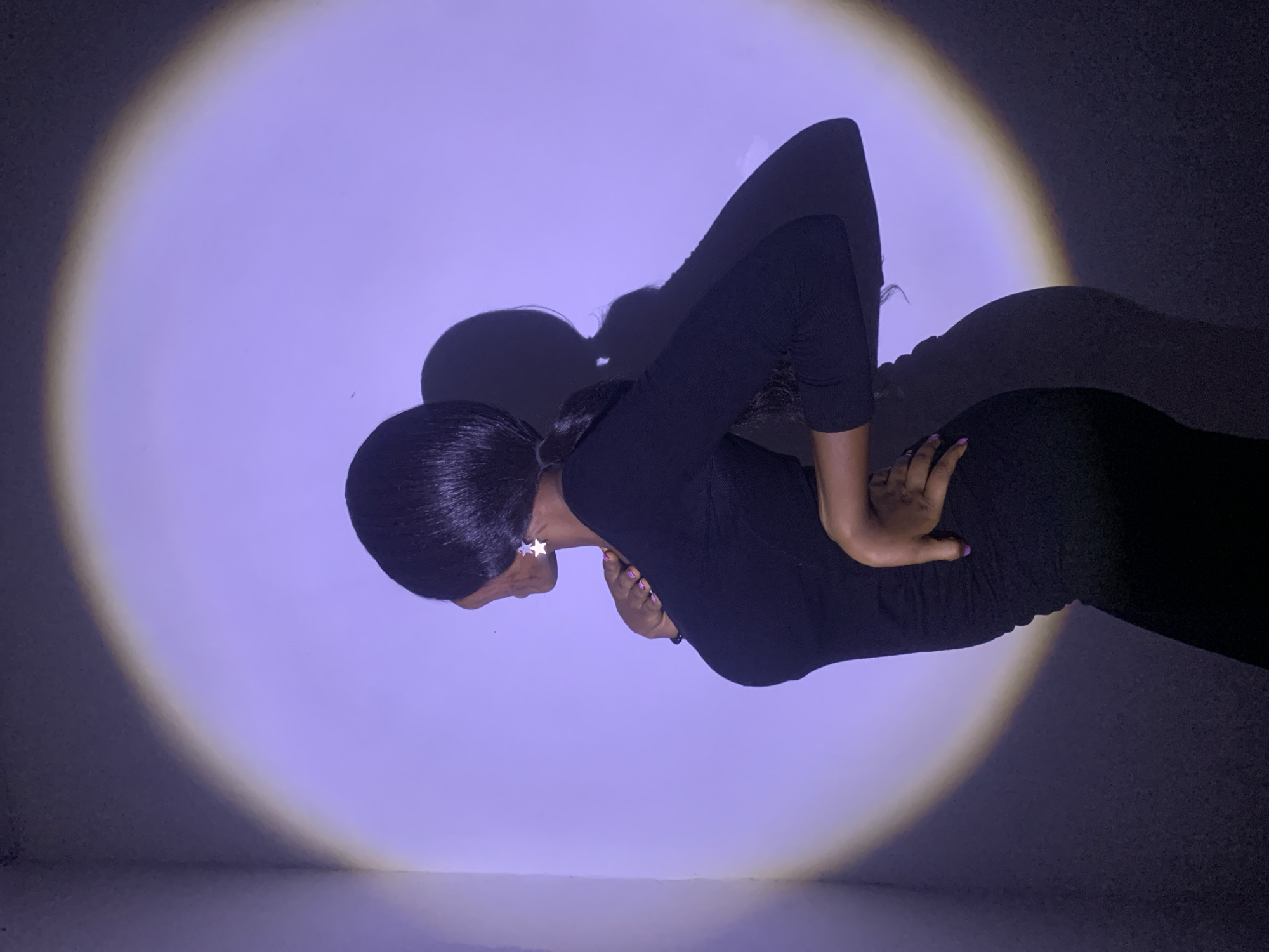 A dark-skinned woman wearing a black, long-sleeved dress stands in a room with a spotlight shining on her. She has short, dark hair, and is wearing star-shaped earrings. Her left hand is on her hip and her right arm is crossed over her abdomen. The walls are a light violet color, and there is a dark shadow cast behind her. The setting is a room interior; any specific location information in Jos, Nigeria, is not apparent from the image.