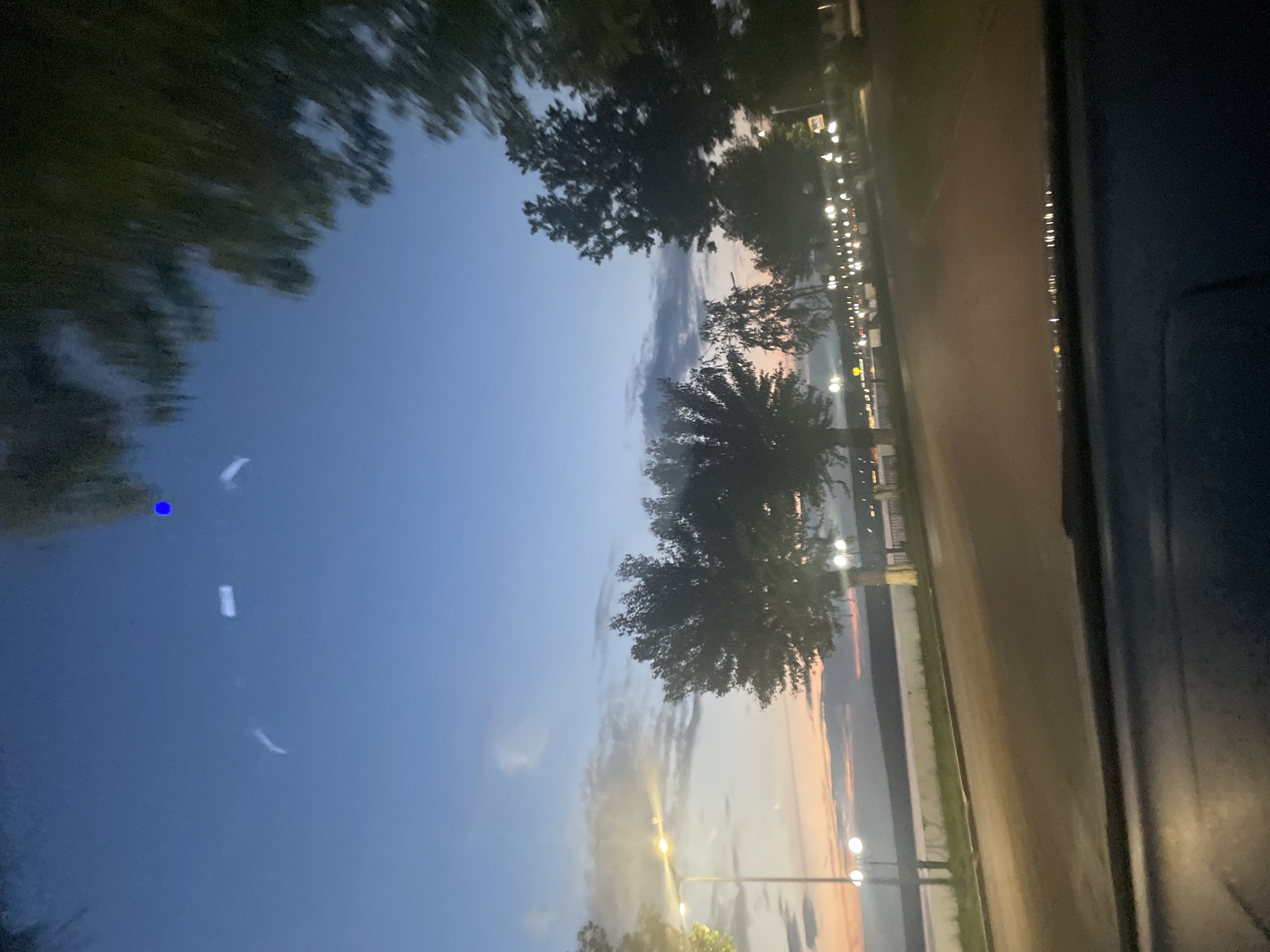 The image captures a twilight scene from inside a vehicle, looking out towards a waterfront promenade. The sky is a gradient of deep blue at the top, transitioning to lighter shades of blue and hints of pink and orange near the horizon, suggesting the sun has recently set. Wispy clouds are visible in the sky.
Along the promenade, there are trees with dark foliage and streetlights emitting a warm glow. A body of water, likely a river or lake, is visible beyond the promenade. The road in the foreground appears to be asphalt, with the interior of the vehicle partially visible at the bottom of the frame.
The location is identified as Votkinsk, Russia. The scene evokes a sense of tranquility and the transition from day to night. There are no people or explicit actions depicted, other than the implied movement of the vehicle. The overall impression is one of a scenic view captured during dusk.