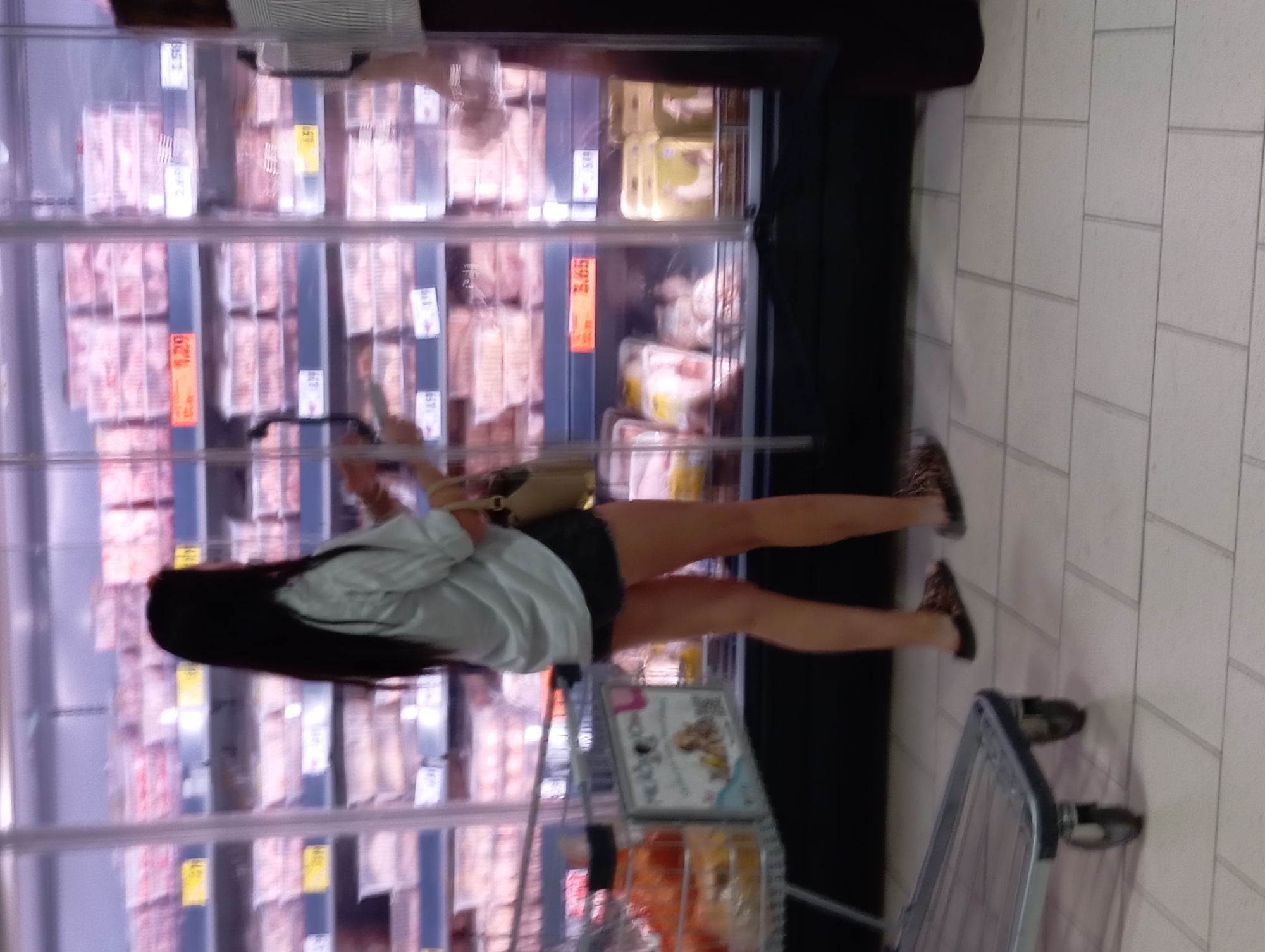 A young woman with long, dark hair, wearing a light-colored shirt and dark shorts, stands in front of a refrigerated display case in a supermarket. She is looking into the case, which is filled with packaged meat products. To her left is a shopping cart with items inside. The floor is tiled in a light color. The supermarket appears to be located in Nitra, Slovakia.