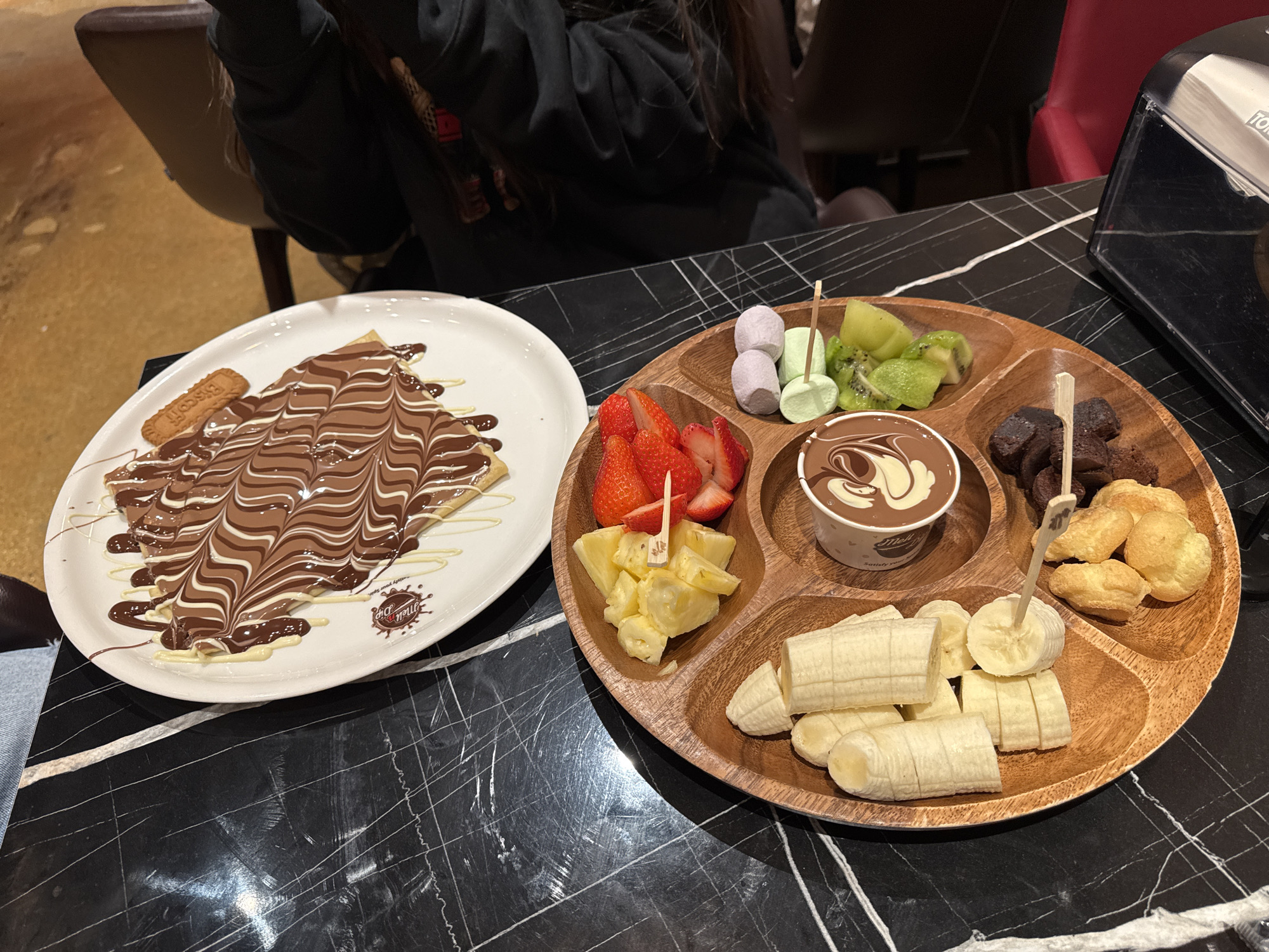 This image depicts a tabletop scene in what appears to be a dessert establishment in Schaumburg, Illinois, United States. The black marble table surface has a white vein pattern. A wooden serving platter containing various fruits, marshmallows, brownies, cream puffs, and a cup of chocolate fondue occupies the right side of the frame. Sliced bananas are most prominent, along with cut strawberries, pineapple chunks, kiwi slices, and multicolored marshmallows. A paper cup filled with swirled chocolate fondue is also on the platter. To the left, there is a white plate topped with a crepe covered in a chocolate and white chocolate drizzle and a Biscoff biscuit. Partially visible are seated individuals, one wearing a black hoodie and another with a light-colored shirt. A tissue dispenser sits on the table.