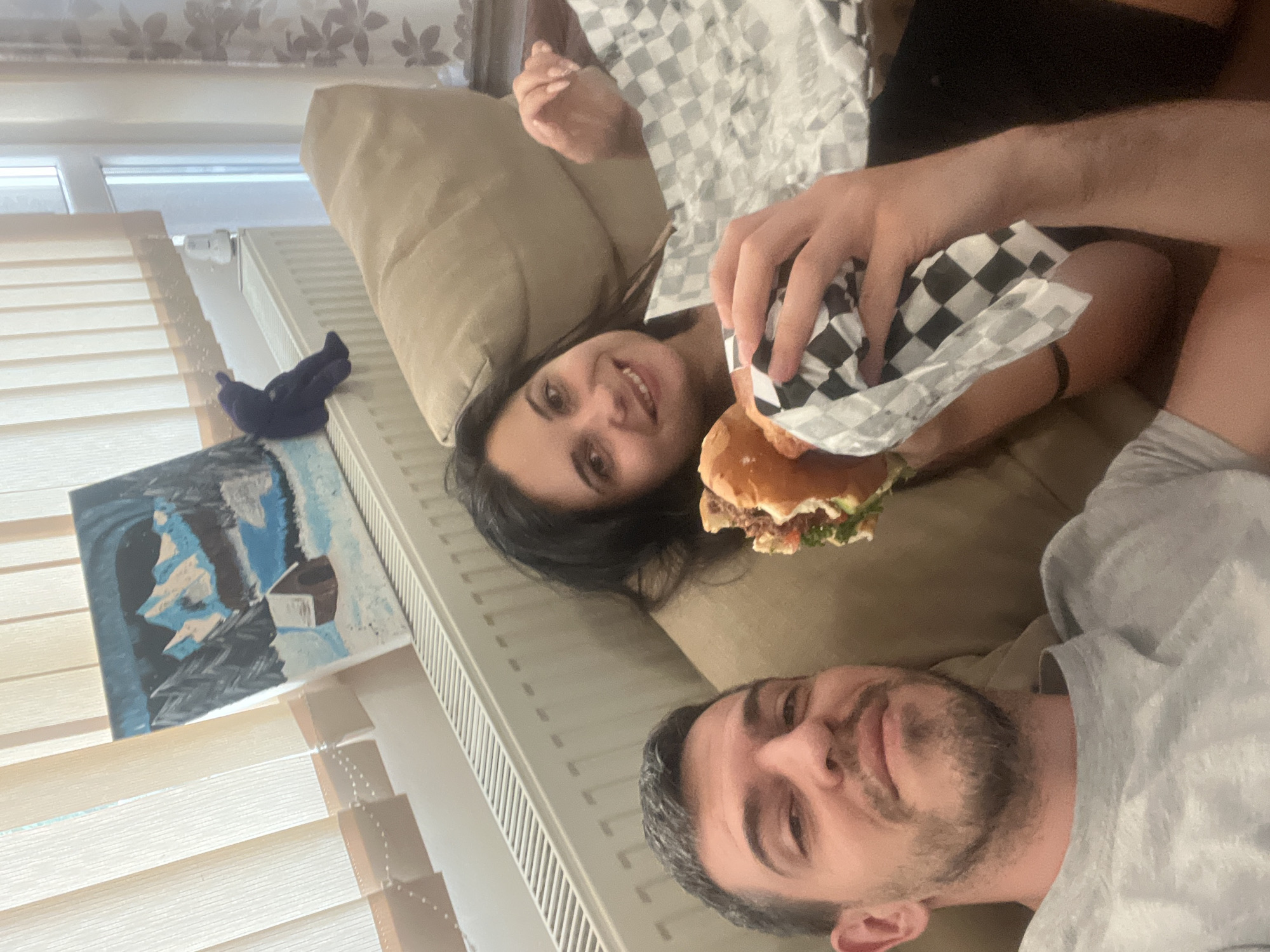 Two individuals, a man and a woman, are lying on a beige sofa in a room with vertical blinds on the left. The man, in the foreground, is wearing a light grey t-shirt and has short, grey hair and stubble. The woman, to his right, has dark hair and is smiling, holding a burger wrapped in checkered paper. She is also wearing a light-colored top. Behind them, a painting of a snowy mountain scene with a small cabin and trees is leaning against a radiator. A small, dark blue plush toy is sitting on top of the radiator next to the painting. The room appears to be an indoor living space, possibly an apartment in Sofia, Bulgaria, given the radiator and blinds which are common in Bulgarian residences.