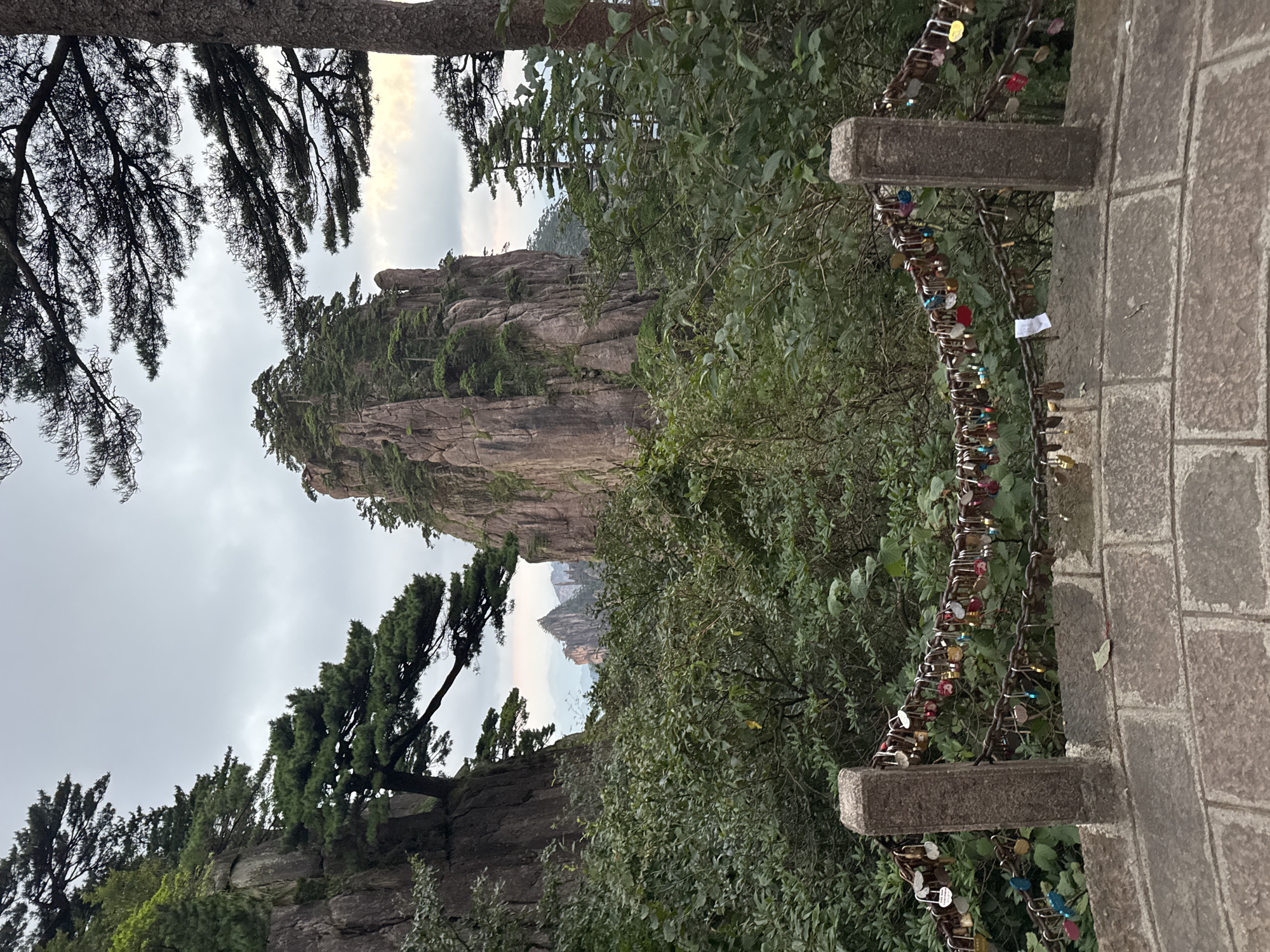 The image depicts a scenic mountain landscape with a prominent rock formation covered in trees in the center. In the foreground, a stone railing is adorned with numerous love locks, attached to chains strung between two stone posts. The railing is constructed of large, rough-hewn stones. Lush green foliage and trees frame the view of the mountains, and a cloudy sky hangs overhead. This appears to be a view from a tourist path in the Huangshan Mountains, known for their distinctive granite peaks and pine trees, located in Huangshan District, China.