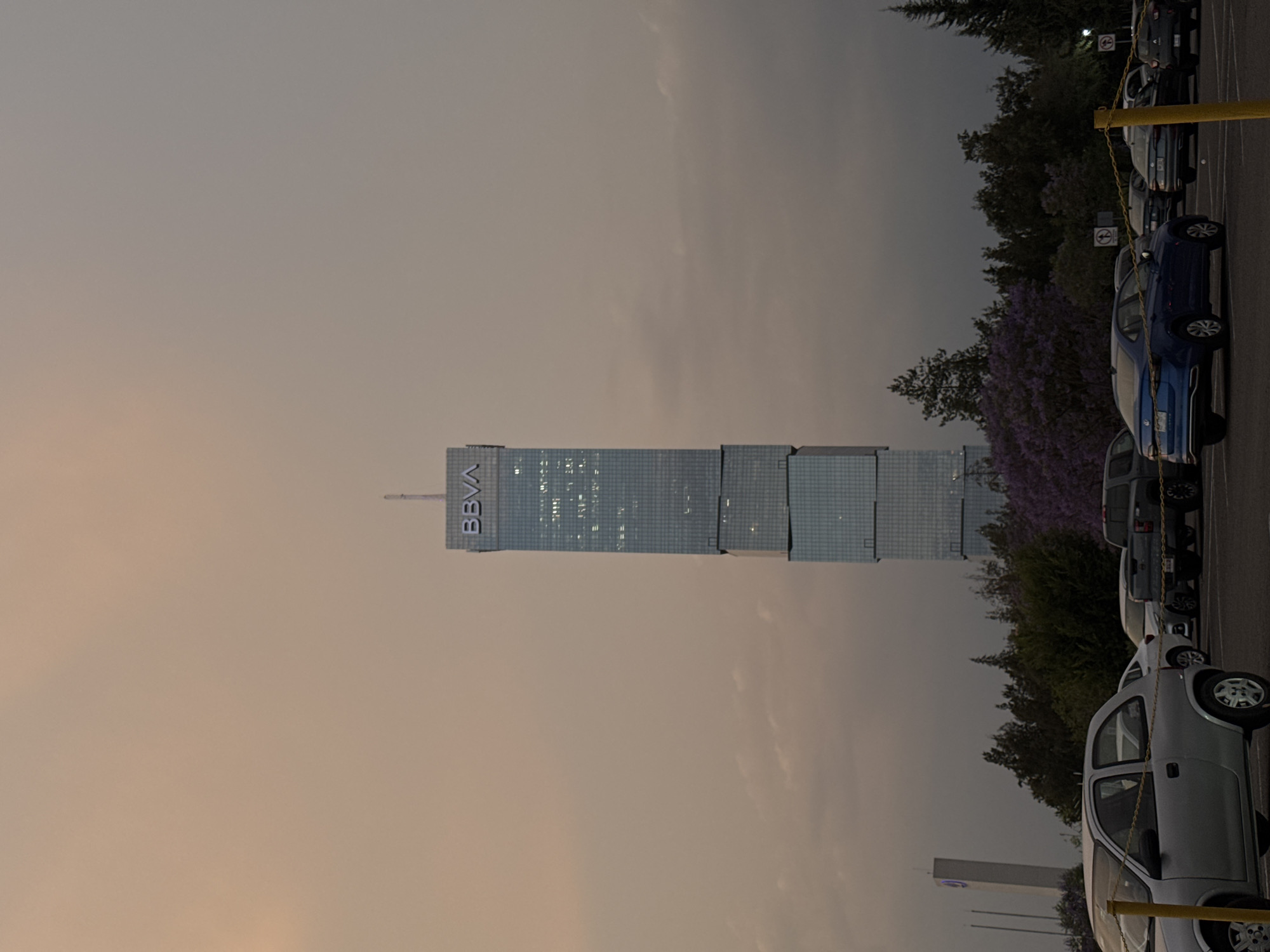 The image shows a parking lot in Heroica Puebla de Zaragoza, Mexico, with several cars parked in marked spaces. A tall, modern skyscraper with the "BBVA" logo is in the background. The sky is overcast, suggesting either early morning or late afternoon. The overall setting is an urban environment on what looks to be the outskirts of the city, as evidenced by the trees and the building in the distance.