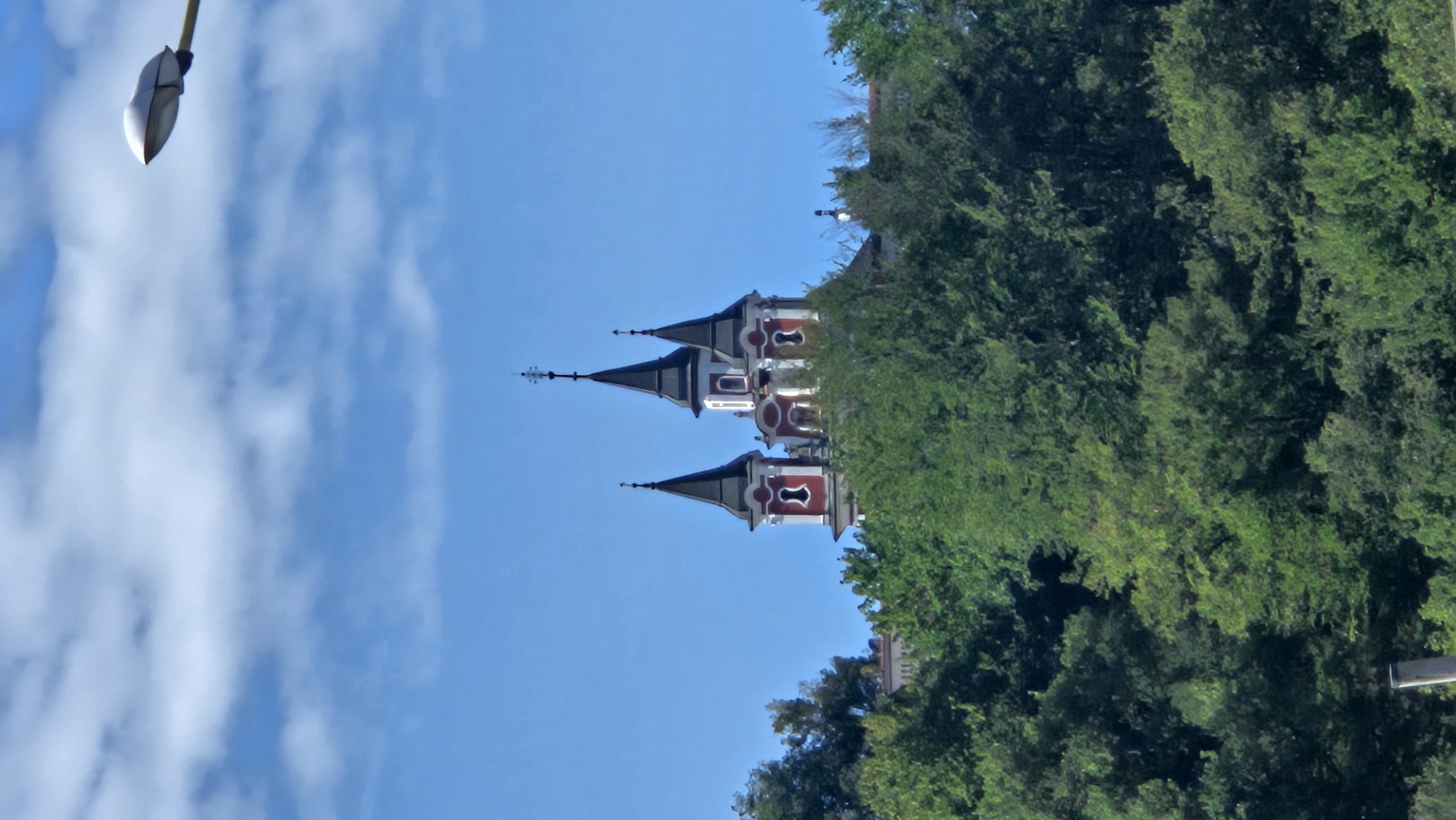 The image shows the Church of the Holy Spirit in Prešov, Slovakia. The church is a prominent building with multiple spires, a red and white facade, and decorative elements. It is situated on a hilltop, partially obscured by lush green trees. The sky is blue with scattered white clouds. In the upper right corner of the image, a modern street lamp is visible, extending into the frame. The church's architectural style suggests a historical religious structure, likely a landmark within the city of Prešov. The surrounding environment is characterized by dense foliage, indicating a natural setting or parkland.