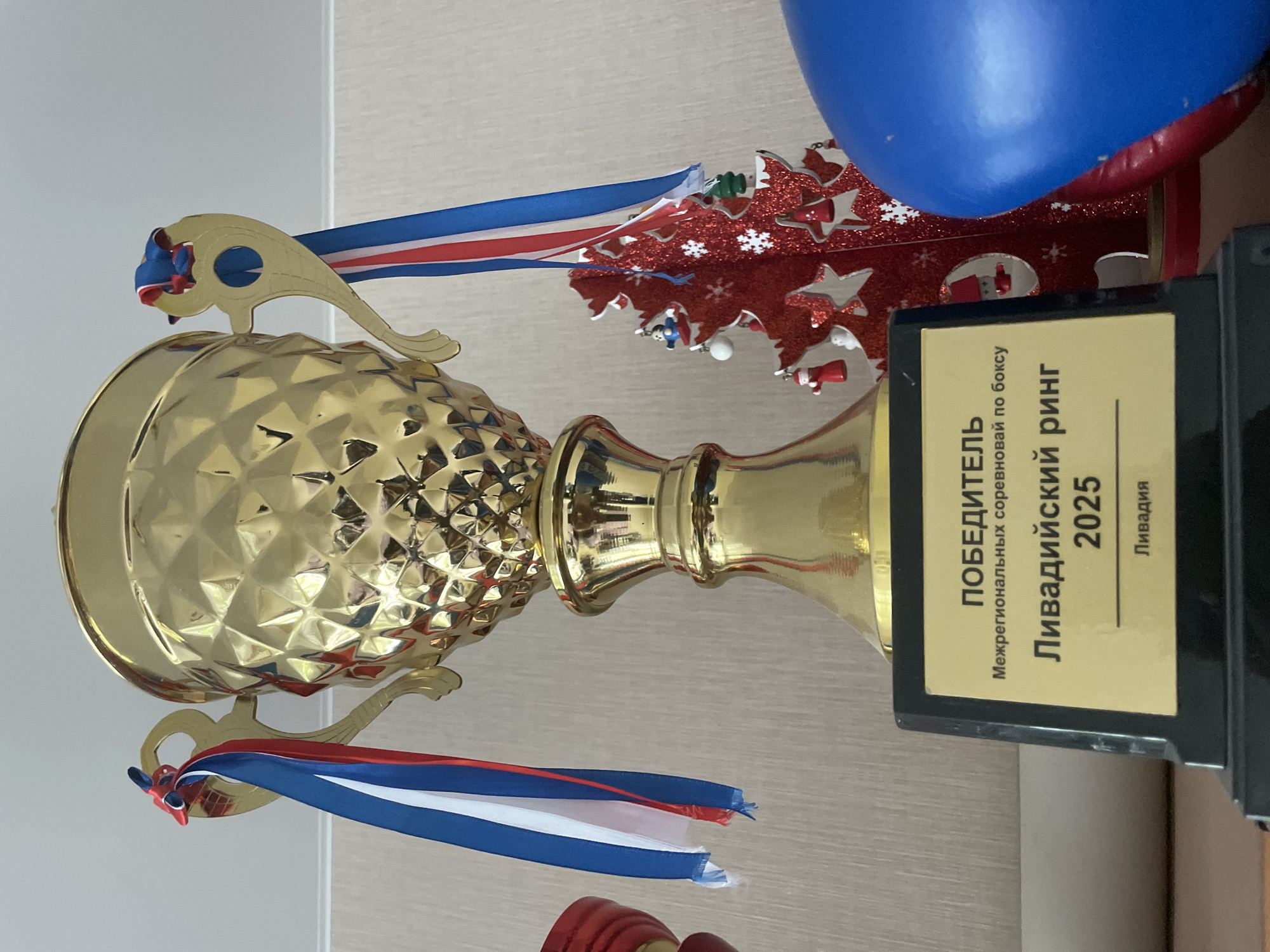 A golden trophy with red, white, and blue ribbons attached to its handles sits on a black base. The base has a gold plate with Russian text that reads "ПОБЕДИТЕЛЬ Межрегиональных соревнований по боксу Ливадийский ринг 2025 Ливадия". To the right of the trophy is a small, red, glittery Christmas tree decoration with small figures and white snowflakes. A portion of a blue inflatable object is visible to the right of the Christmas tree. The background appears to be a wall with a textured wallpaper pattern.