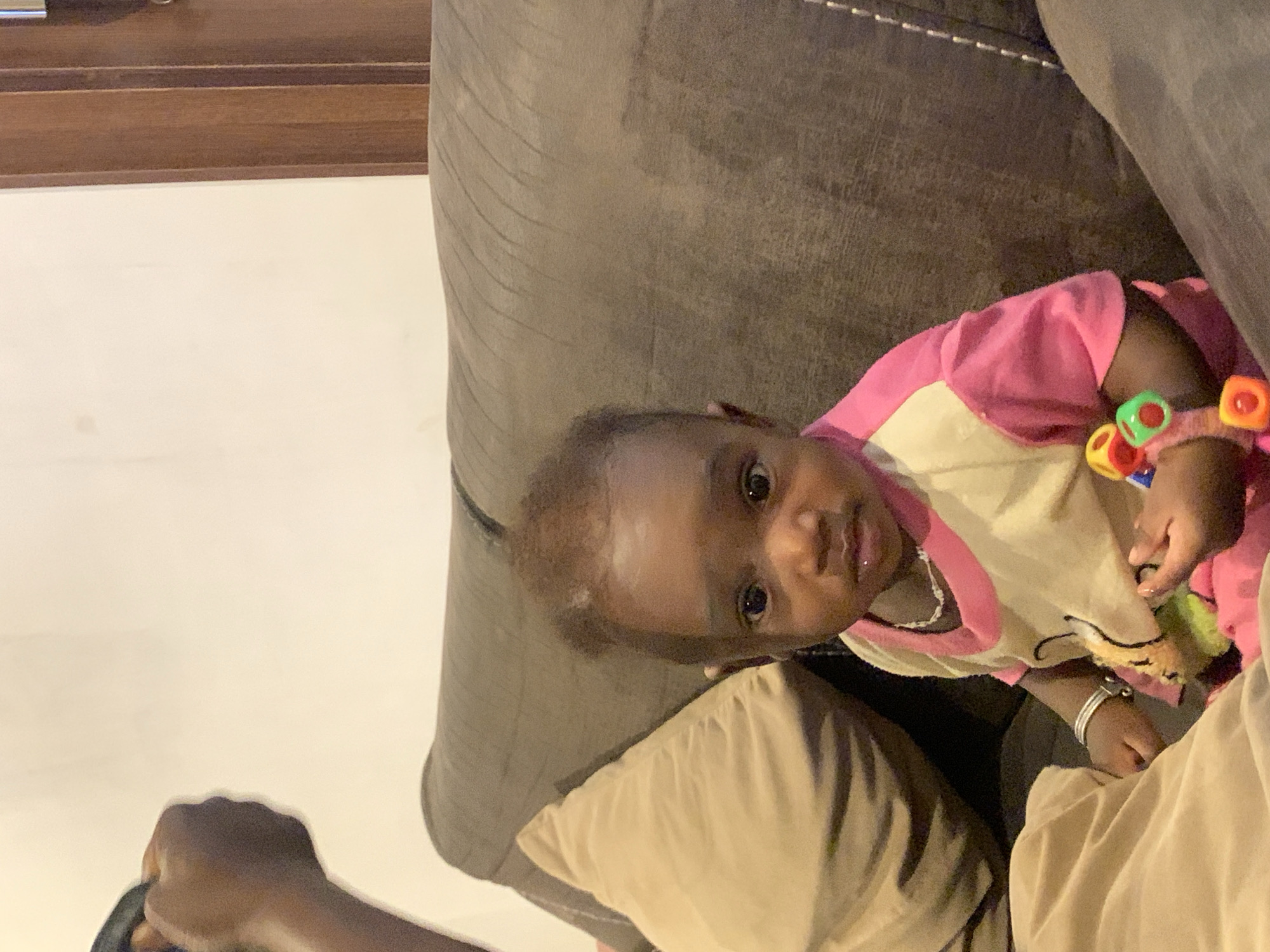 A baby with brown skin, wearing a pink and tan shirt and a toy bracelet with colorful beads, sits on a brown couch. The baby is looking at the camera. An arm and hand of a person with brown skin holding a dark object are visible in the lower left corner. The setting appears to be indoors with a white wall and a brown trim visible on the left. The scene is speculated to be set in Jos, Nigeria.