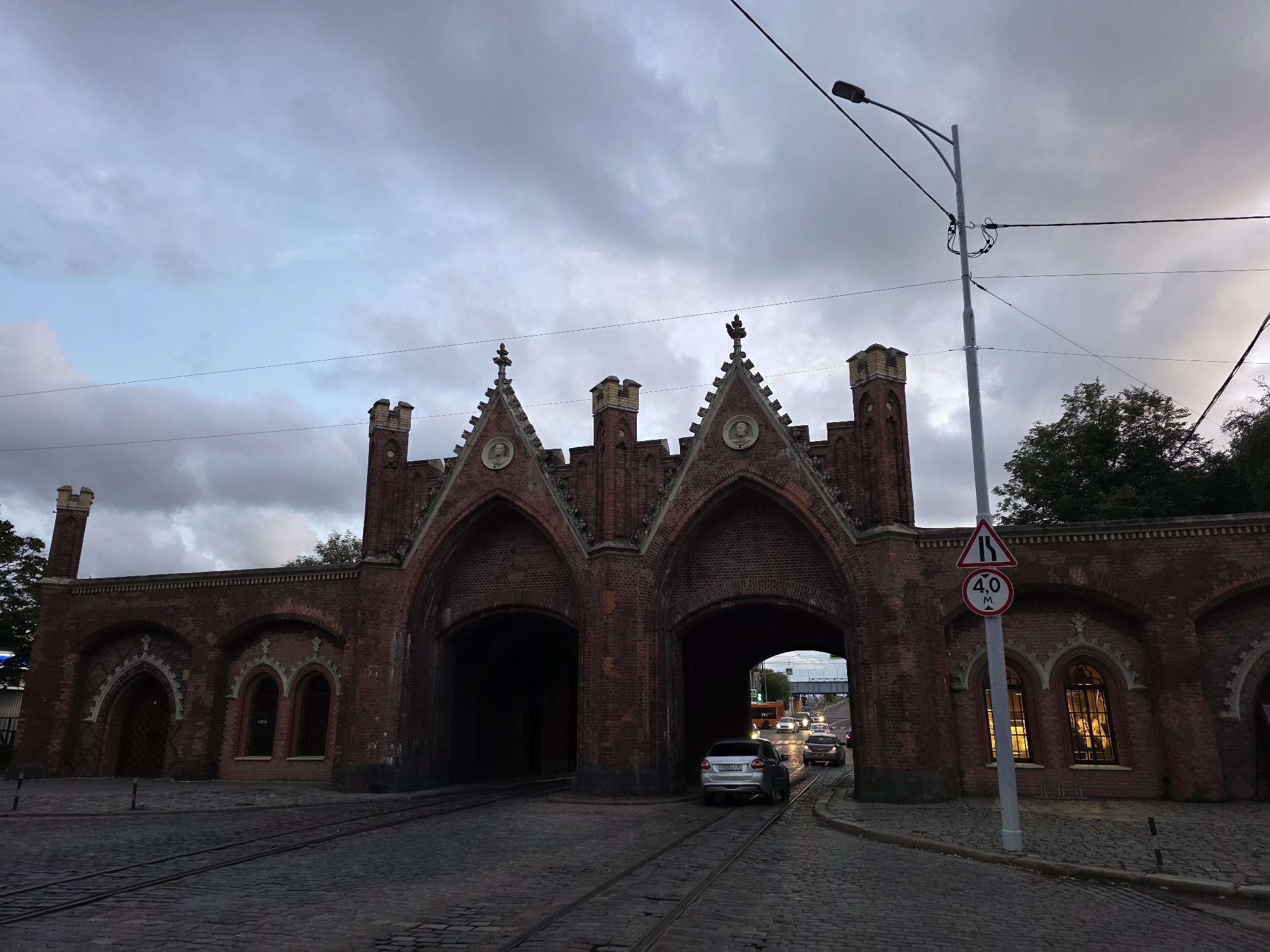 The image captures a frontal view of the Brandenburg Gate in Kaliningrad, Russia. The gate, constructed from red brick, features two arched passages, Gothic architectural elements, and decorative spires. Paved cobblestone streets with visible tram tracks lead towards the gate. Several cars are seen passing through the right archway, with one silver sedan positioned prominently. A street light with multiple wires is visible on the right side of the frame, along with traffic signs, including a triangular sign indicating a narrow road and a circular sign with a "4.0 M" inscription, likely denoting a height restriction. The sky is overcast with dark clouds, and the windows of the gate's flanking structures are illuminated from within. The scene appears to be set in the late afternoon or early evening.