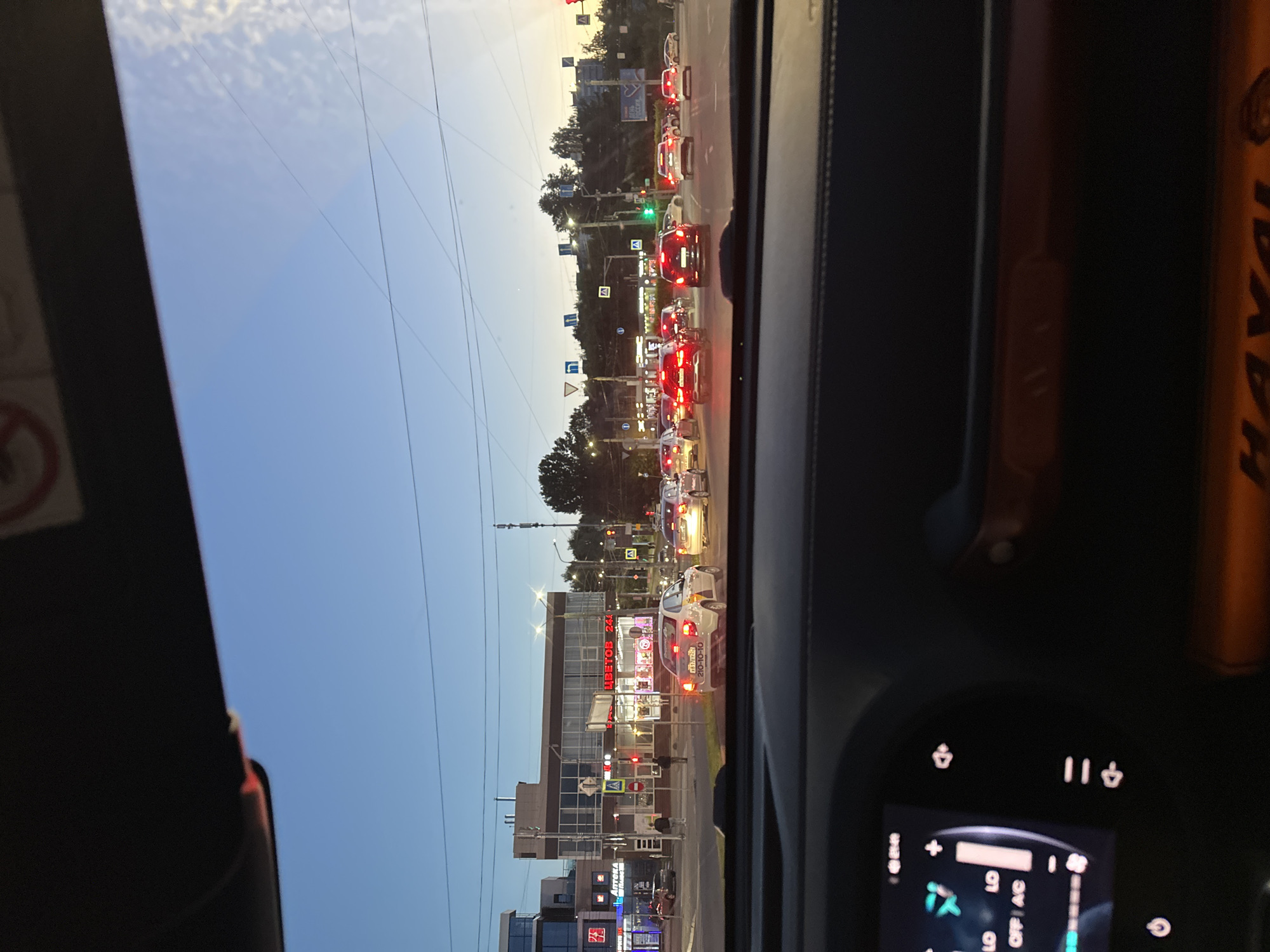 This is a slightly tilted, vertical, eye-level, outdoor shot taken from the perspective of someone inside a car, looking through the windshield. The scene depicts a street in Nizhny Novgorod, Russia, during what appears to be early evening or late afternoon. The sky is a clear, pale blue with some wispy clouds and hints of the setting sun.
The main subject is a street scene with moderate traffic. Cars are lined up, indicating a potential traffic jam or slow-moving traffic. Red brake lights are visible on many vehicles. A traffic light is also present and appears to be green, suggesting the traffic may be about to move. Along the street, there are buildings, trees, and streetlights. One building prominently features a sign with Cyrillic text, including "ЦВЕТОВ 24ч" which translates to "FLOWERS 24h". Another building has "АПТЕКА" visible, meaning "PHARMACY". There are also various street signs, some with traffic symbols and others with directions.
The interior of the car is partially visible on the right side of the frame, showing a dashboard with a digital display. The display shows various icons and numbers, including what looks like a temperature reading, a gear indicator ("D"), and possibly media controls. A small portion of an orange object with "HAVAL" written on it is also visible on the right, likely part of the car's interior trim or a piece of equipment.
The overall atmosphere is calm and ordinary, depicting a typical urban scene at dusk. The lighting suggests natural daylight is fading. No people are clearly discernible in the scene, though silhouettes in cars and on the sidewalk might be present but are not distinct.