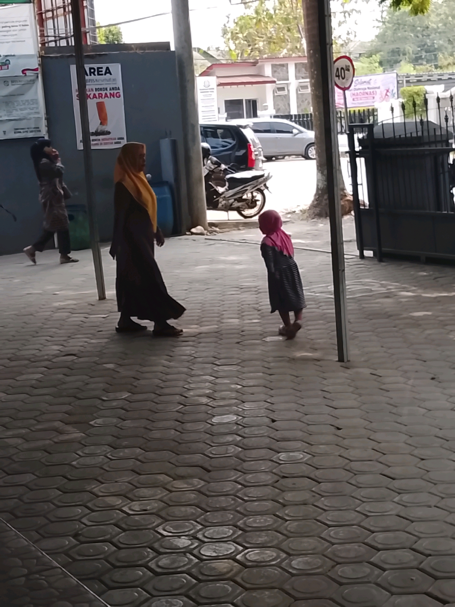 In the city of Slawi, Indonesia, a paved sidewalk with hexagonal tiles leads to a building entrance. A sign on the wall reads "AREA DILARANG MEROKOK SEKARANG," which translates to "SMOKING AREA NOW PROHIBITED." To the left, a person in dark clothing walks away from the viewer, and to the right, a woman in a dark abaya and a yellow headscarf is walking. Further to the right, a small child wearing a pink hijab and a striped dress walks away from the viewer. A speed limit sign indicating "40" is visible on a pole, along with banners in the background. A parked motorcycle is visible to the left.