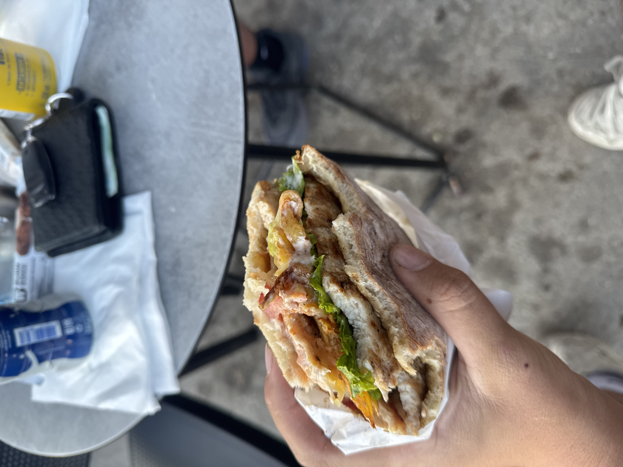 The image displays a close-up, high-angle view of a person's hand holding a large, hearty sandwich in an outdoor setting. The sandwich features a toasted or grilled flatbread, possibly ciabatta, packed with layers of grilled chicken or meat strips, crisp green lettuce, and what appear to be golden-brown potato strips or fries, all bound together with a creamy white sauce. It is partially wrapped in white paper, indicating it's a takeaway or street food item.
The scene unfolds on a patio or outdoor dining area, with a light gray, textured round table positioned to the left. On the table, several personal items are visible: a black wallet or cardholder with a key attached, a partially obscured yellow bottle with black text (possibly "VITA"), a blue and white bottle with a barcode and some unreadable white text, and crumpled white paper or napkins. The background is blurred, revealing dark metal furniture legs and a mottled gray concrete ground. A white sneaker is partially visible in the lower right, suggesting someone's foot resting on the ground.
The activity depicted is the casual enjoyment of a meal outdoors. The bright lighting suggests it is daytime, likely sunny. While no specific location cues are visible beyond the general outdoor setting, the activity is consistent with enjoying street food in a city like Plovdiv, Bulgaria. No emotions are discernible from the partial view of the person, but the scene evokes a sense of a quick, satisfying meal.