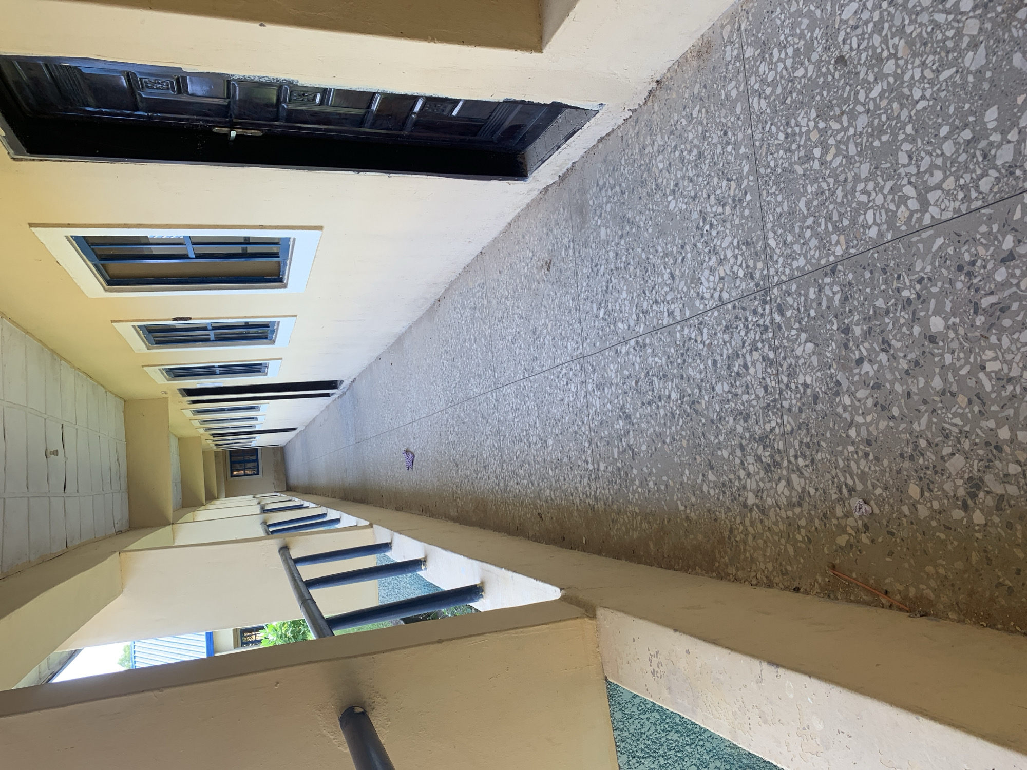 The image depicts an exterior hallway of a building, likely an educational institution or government office, located in Jalingo, Nigeria. The hallway is paved with terrazzo flooring, a common material in such environments. It is flanked by a cream-colored wall on the right, featuring multiple dark-colored doors and windows with blue frames. The left side of the hallway is an open-air walkway with a railing, leading to more rooms. The overall impression is one of a functional, possibly older, building, showing signs of wear and tear. A crumpled piece of cloth lies on the floor near the center of the hallway, and a thin, orange stick is visible near the wall on the left. The ceiling of the hallway appears to be suspended, with visible acoustic tiles. In the background, through a window, a glimpse of lush green vegetation and another building with a blue roof suggests the surrounding environment is tropical.