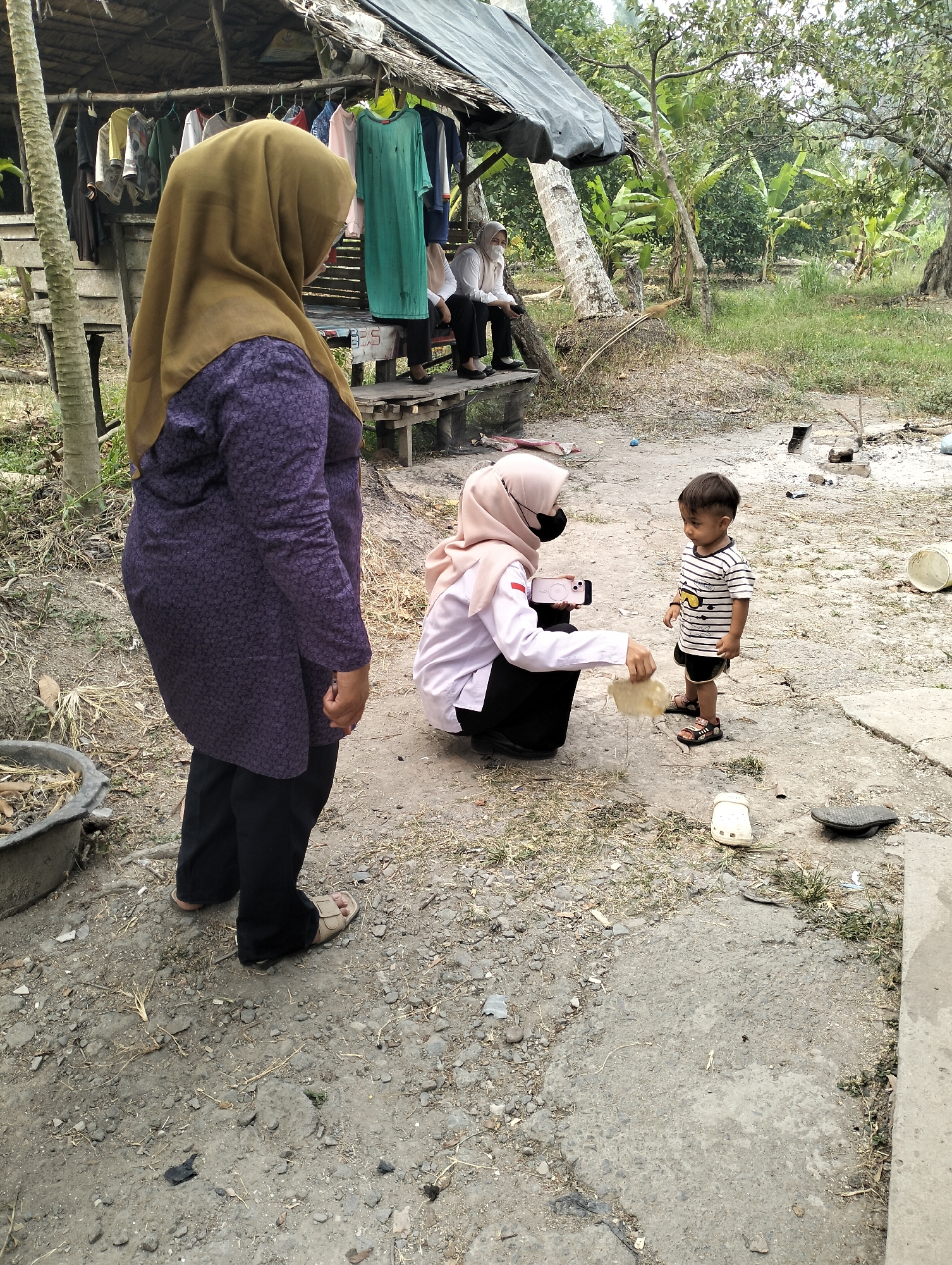 In Arisan Gading, Indonesia, a scene unfolds under a makeshift, tarp-covered shelter with a thatched roof. The shelter displays a rack of various garments, including a bright green dress and t-shirts. To the right, a young child, wearing a striped t-shirt and shorts, stands on a dirt and gravel ground. A woman, dressed in a light pink hijab and a white collared shirt with a red stripe on the sleeve, kneels in front of the child, holding a white phone and a light-colored piece of food or material. Another woman, in a brown hijab and a purple patterned shirt, stands to the left, facing away from the camera. In the background, lush green foliage and trees suggest a rural or semi-rural setting. Scattered on the ground are various objects, including sandals, a plastic bucket, and what appear to be remnants of a fire pit. The overall atmosphere is casual and appears to be a warm, outdoor environment.
