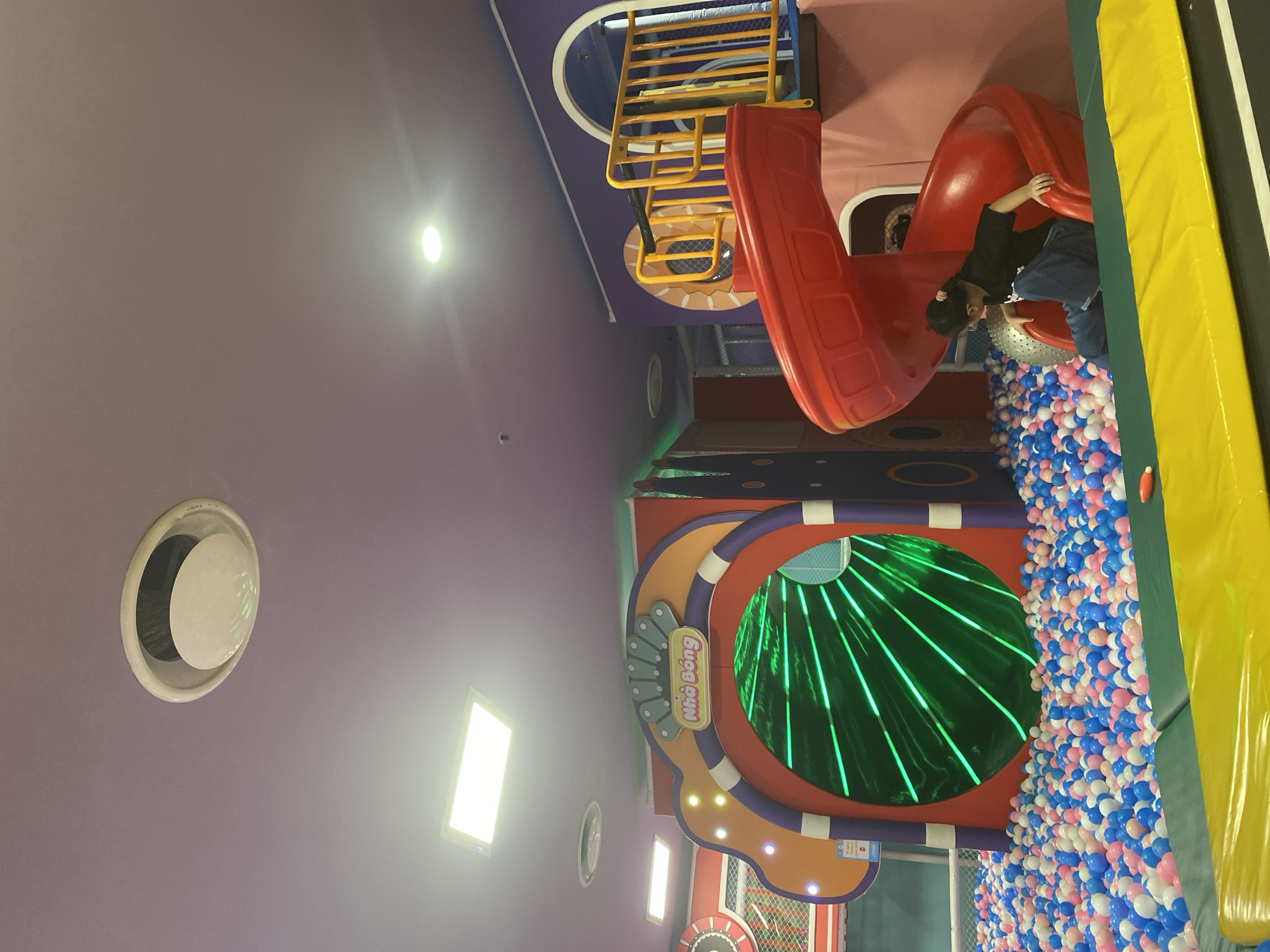 A young girl, wearing a black t-shirt and blue jeans, is captured mid-action on a red spiral slide, her hands gripping the edge as she descends. She is situated within a vibrant indoor play area, likely at "Nhà Bông" in the city of Phu Thuy, Vietnam. The play area is filled with a colorful sea of blue, pink, and white balls, creating a soft landing zone. To the left, a large, tunnel-like entrance glows with green neon lights, adding a futuristic and engaging element to the space. The ceiling, a soft purple hue, is dotted with circular and rectangular lights, illuminating the playful environment. The background reveals more of the colorful, imaginative play structures, including yellow railings and purple walls adorned with circular motifs, suggesting a playful and stimulating atmosphere designed for children's entertainment.