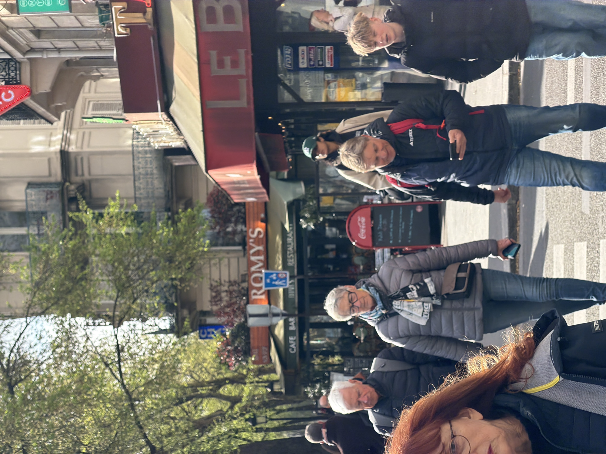 Here is a detailed description of the image:
The image captures a bustling street scene in Paris, France. Several pedestrians are visible, including:
* A younger man with blonde hair wearing a black jacket and jeans
* A man in a black jacket with a red backpack and blue jeans, seemingly using a phone.
* An older woman with gray hair, glasses, a gray puffy jacket, a scarf with a money print, and blue jeans is also holding a phone.
* A couple of older individuals in dark jackets and what appears to be the back of an older woman with red hair and glasses, situated in the foreground.
The scene takes place in front of a building with red awning signs, reading "LE...". On the same side of the street, farther down, a restaurant called "ROMY'S RESTAURANT" is visible. A Coca-Cola sign and a menu board are outside of Romy's.
The environment includes trees with green leaves and glimpses of other buildings. The overall impression is a casual street shot, capturing a normal day in Paris.